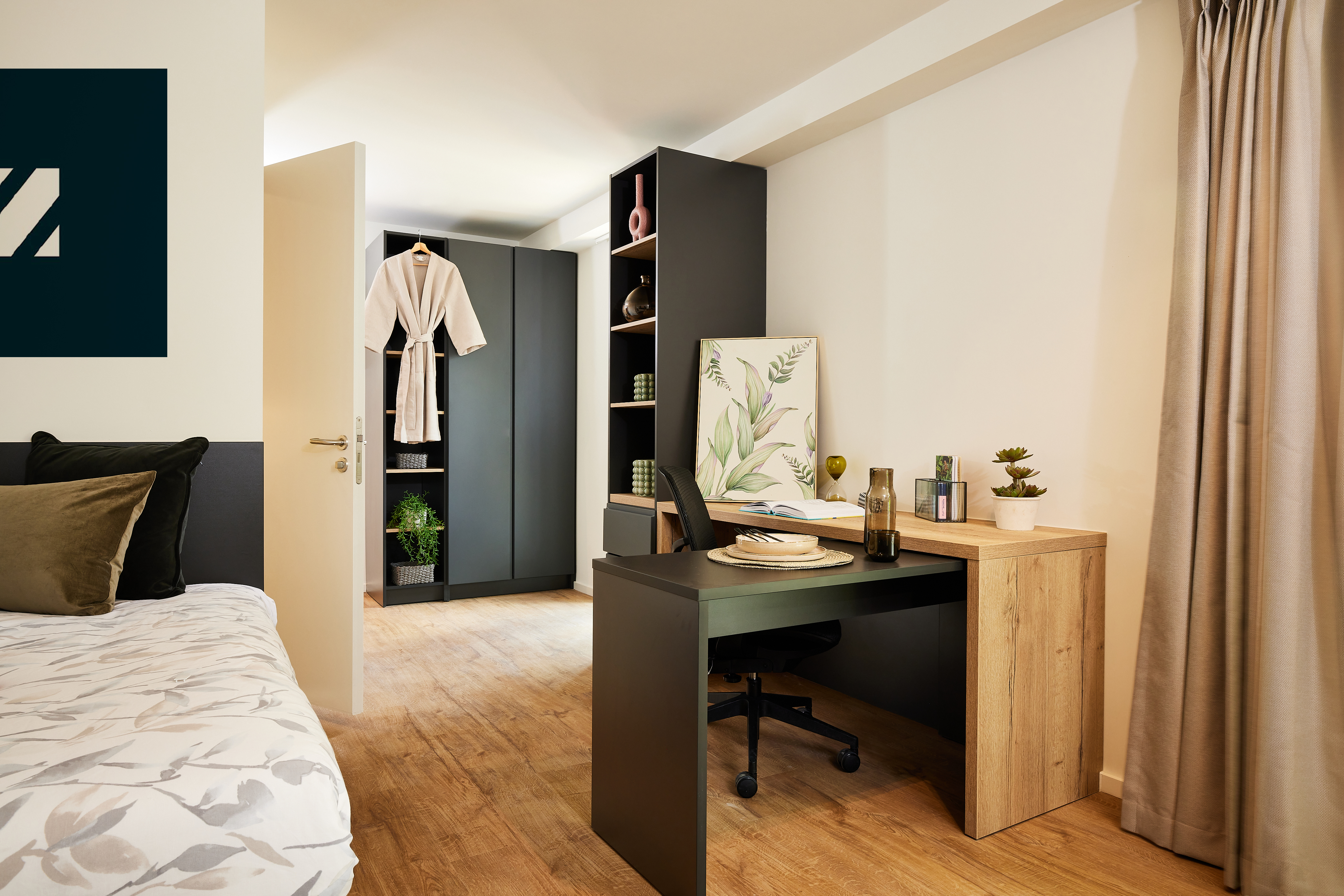 A modern bedroom with a small office space. A bed with white patterned linens rests beside a wooden desk adorned with a laptop and decor. A robe hangs in an open wardrobe, while natural light bathes the room through a window, highlighting elements reminiscent of sleek copper architecture.