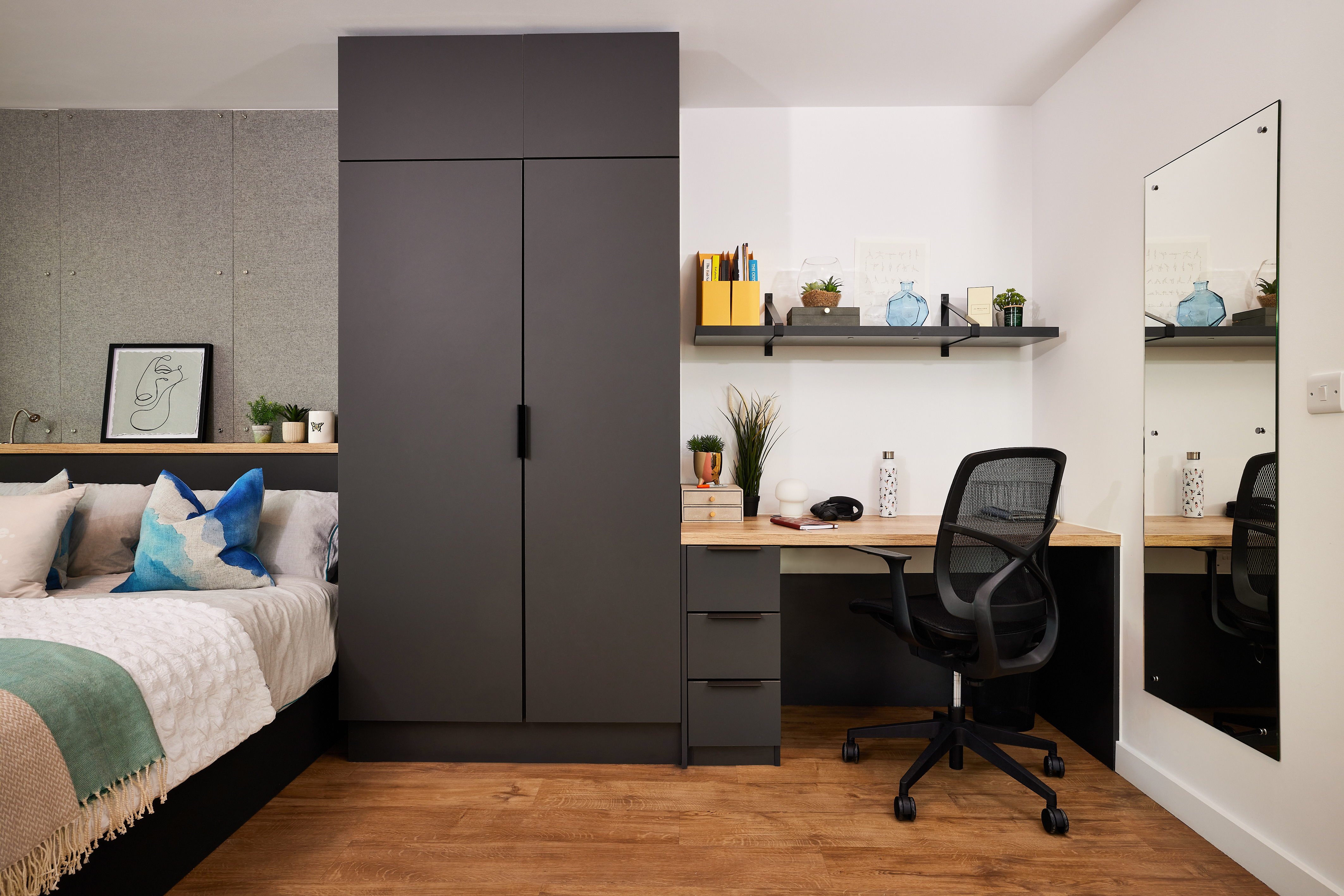 A modern bedroom at Lewisham Exchange features a large dark wardrobe, a wooden desk with drawers, and a black office chair. The wall showcases a shelf with decorative items, while the bed with pillows rests on the left. A tall mirror stands on the right, complementing the elegant wooden floor.