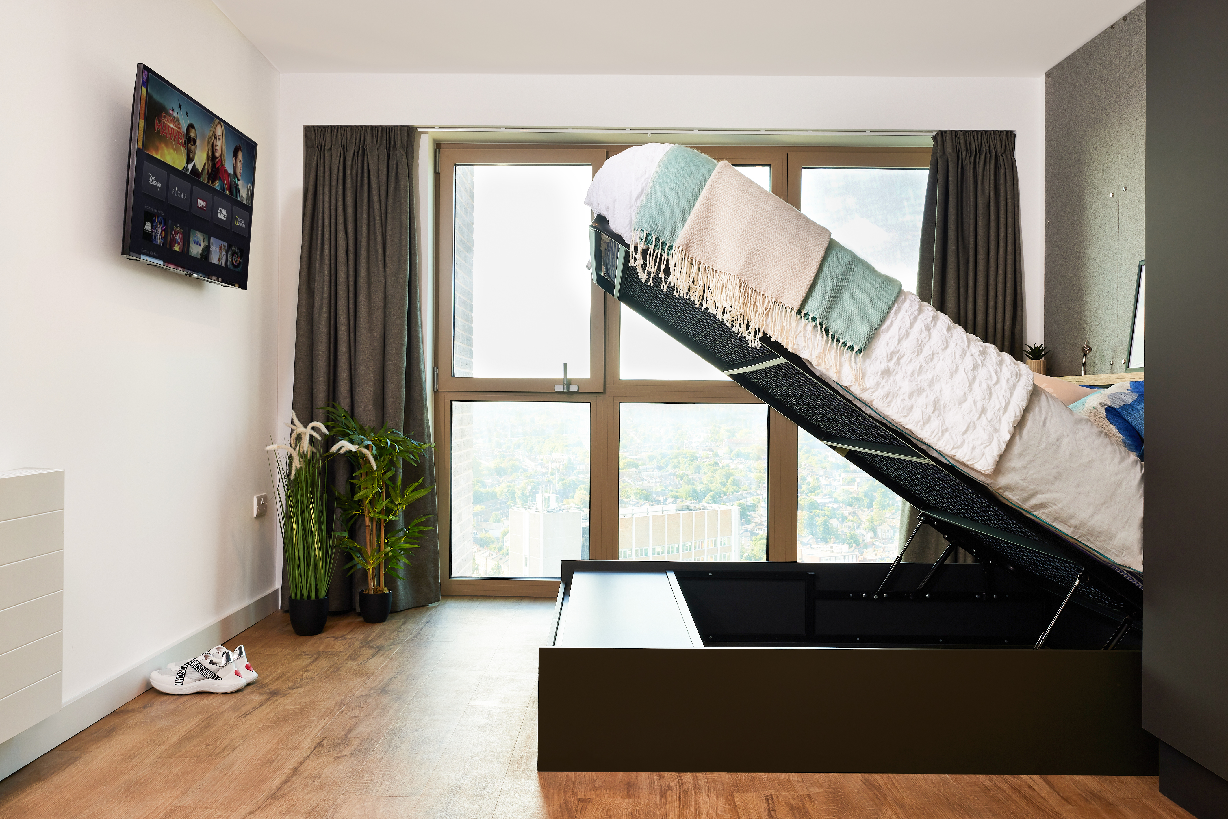 The bright room at Lewisham Exchange features a bed in an upward, open storage position. A TV on the wall displays a streaming platform. Large windows with closed gray curtains overlook the view. White sneakers and a potted plant sit near the radiator on the wooden floor.