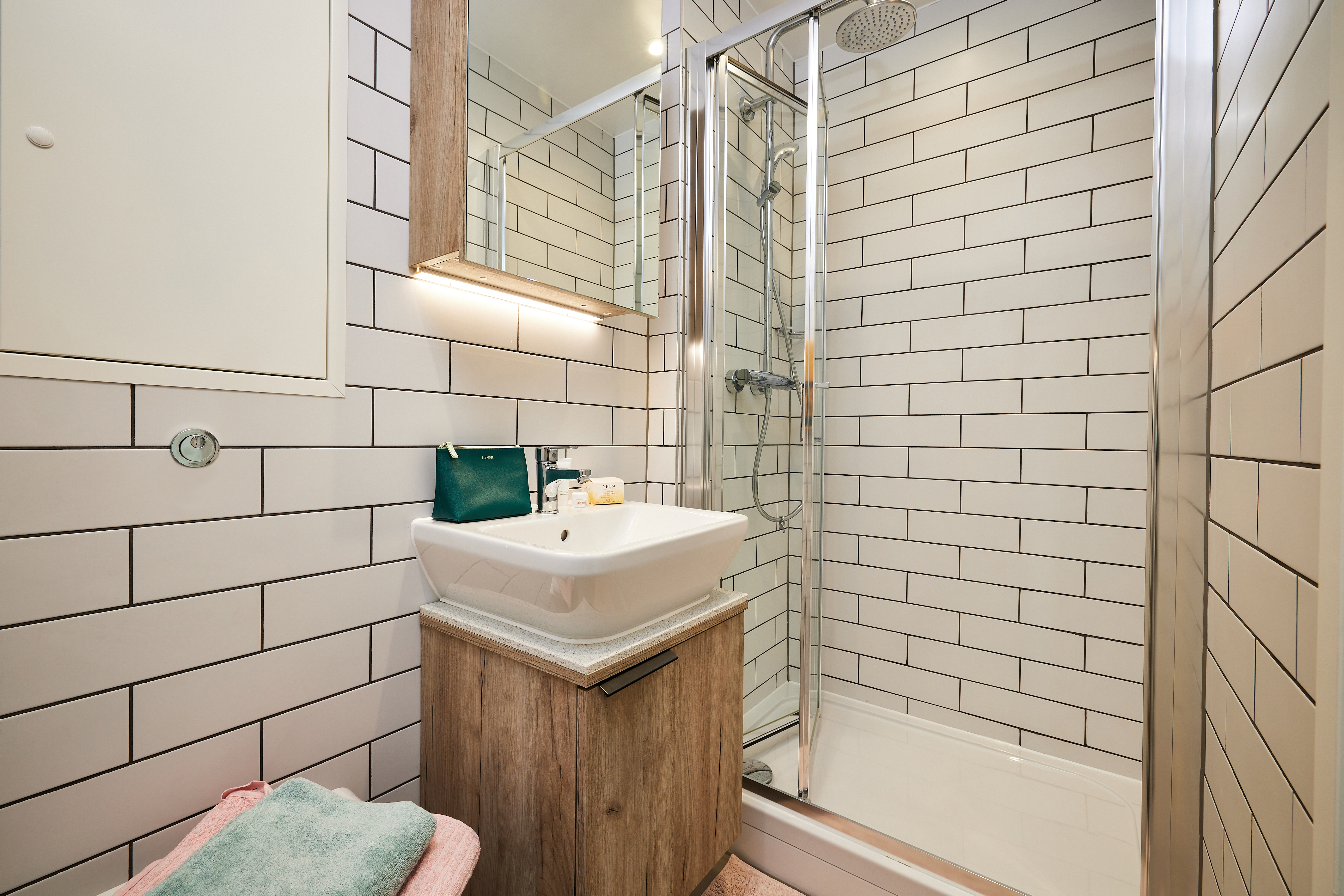 This compact bathroom in Lewisham Exchange features a white subway tile shower with a glass door and rainfall showerhead. A small wood-accented sink with cabinet sits below a mirror, while a pink towel is neatly folded on a nearby surface.