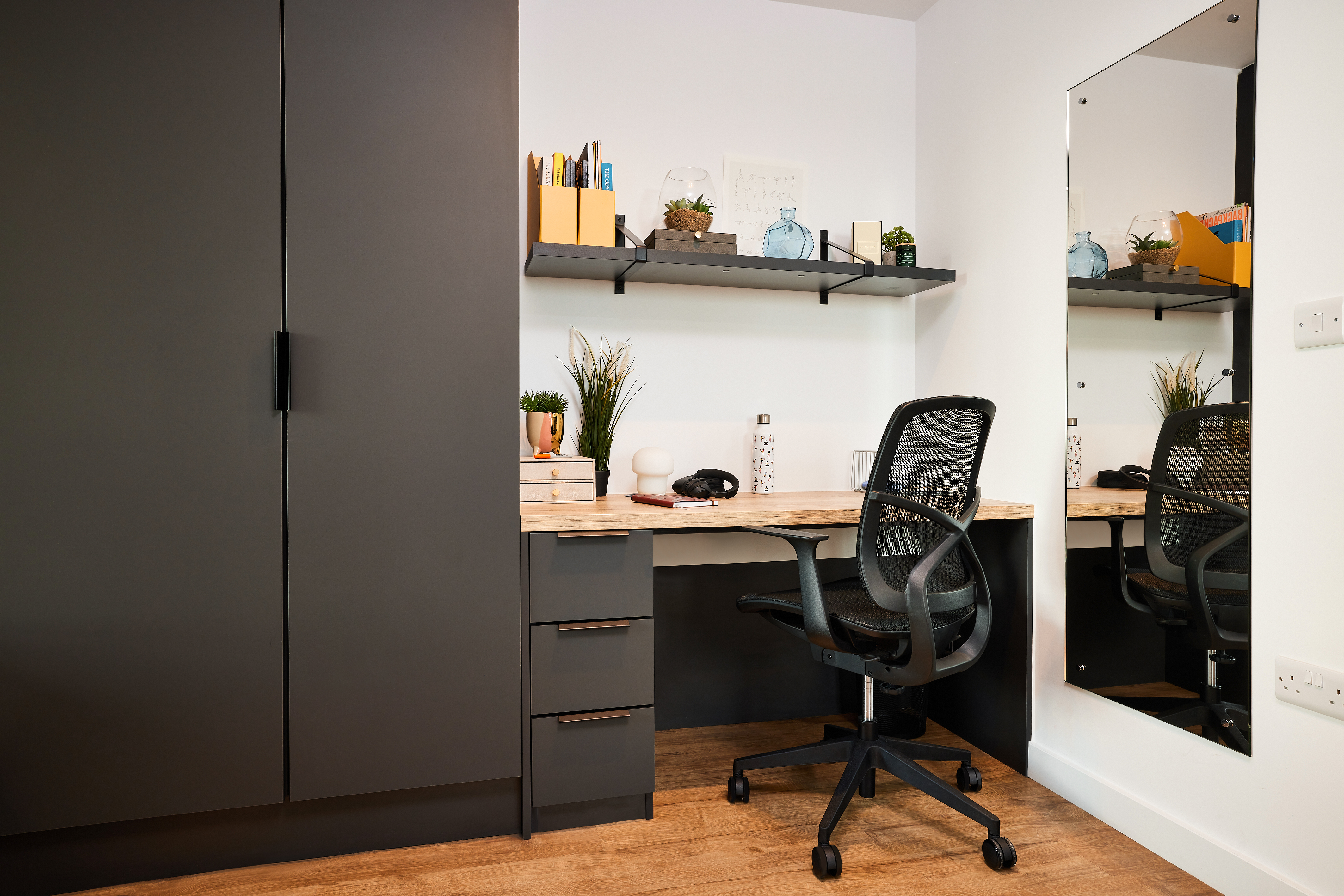 The minimalist home office at Lewisham Exchange features a wooden desk, black mesh office chair, and gray filing cabinets. A shelf above the desk holds books, plants, and decorative items. A large mirror on the adjacent white wall reflects part of the serene room.