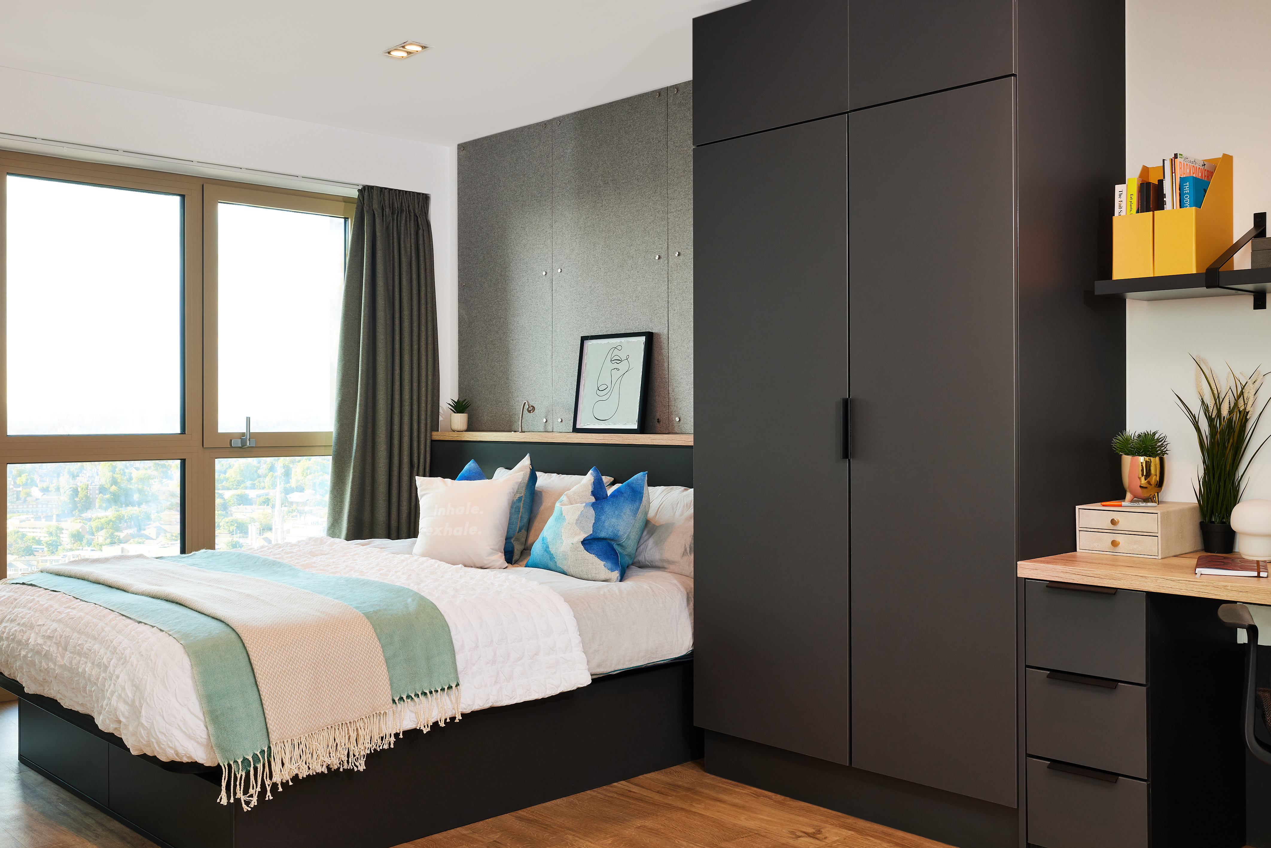 The modern bedroom at Lewisham Exchange boasts a large bed with white and blue pillows and a light throw blanket. A sleek black wardrobe beside a desk with shelves complements the space, while a large window with curtains allows natural light to highlight the artwork above the bed.