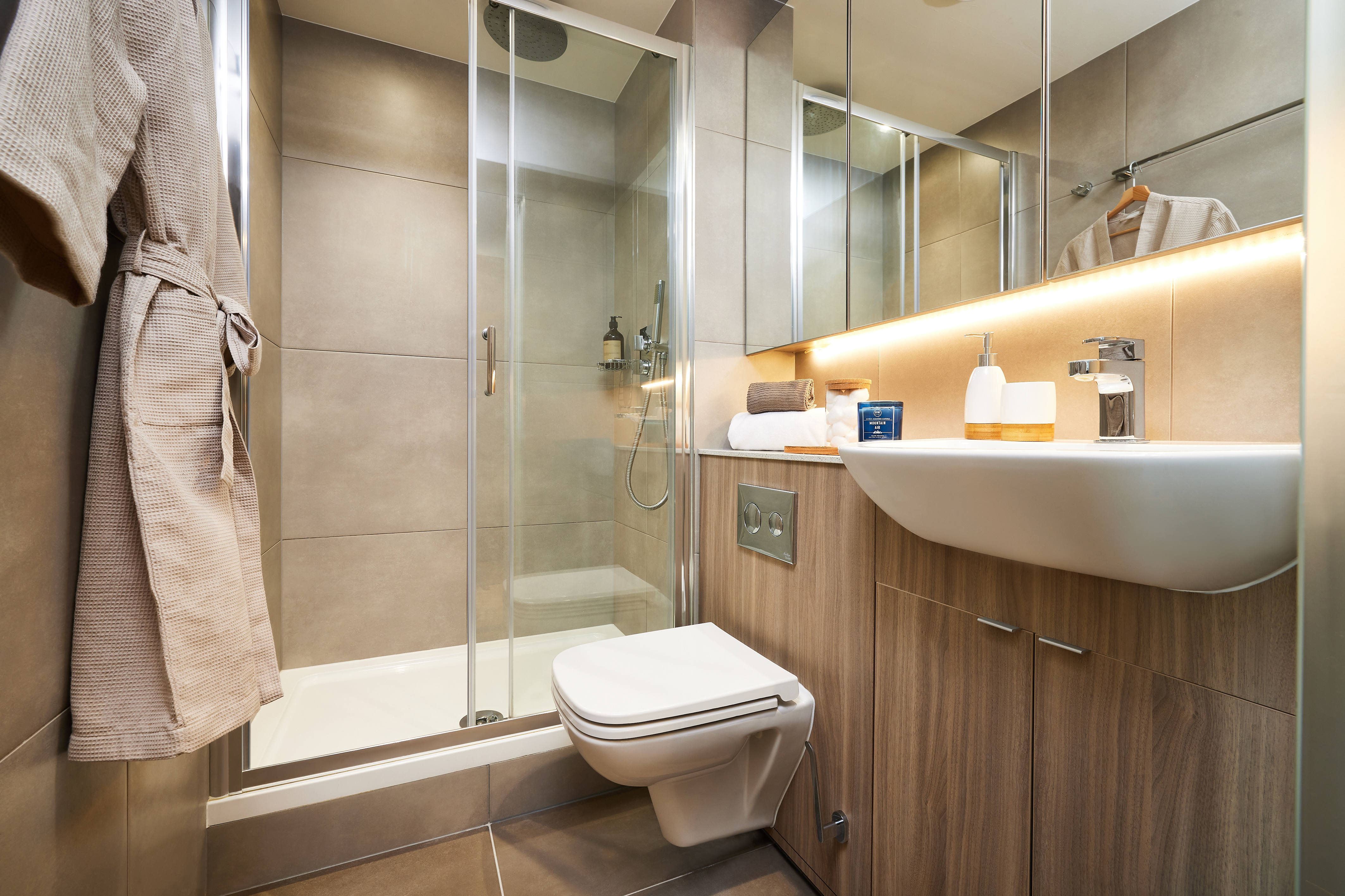 Modern bathroom at Strawberry Place with a glass-enclosed shower, wall-mounted toilet, and a sink featuring wooden cabinetry. Towels and toiletries are neatly arranged on the counter. Warm lighting highlights the sleek, neutral-toned tiles and fixtures.
