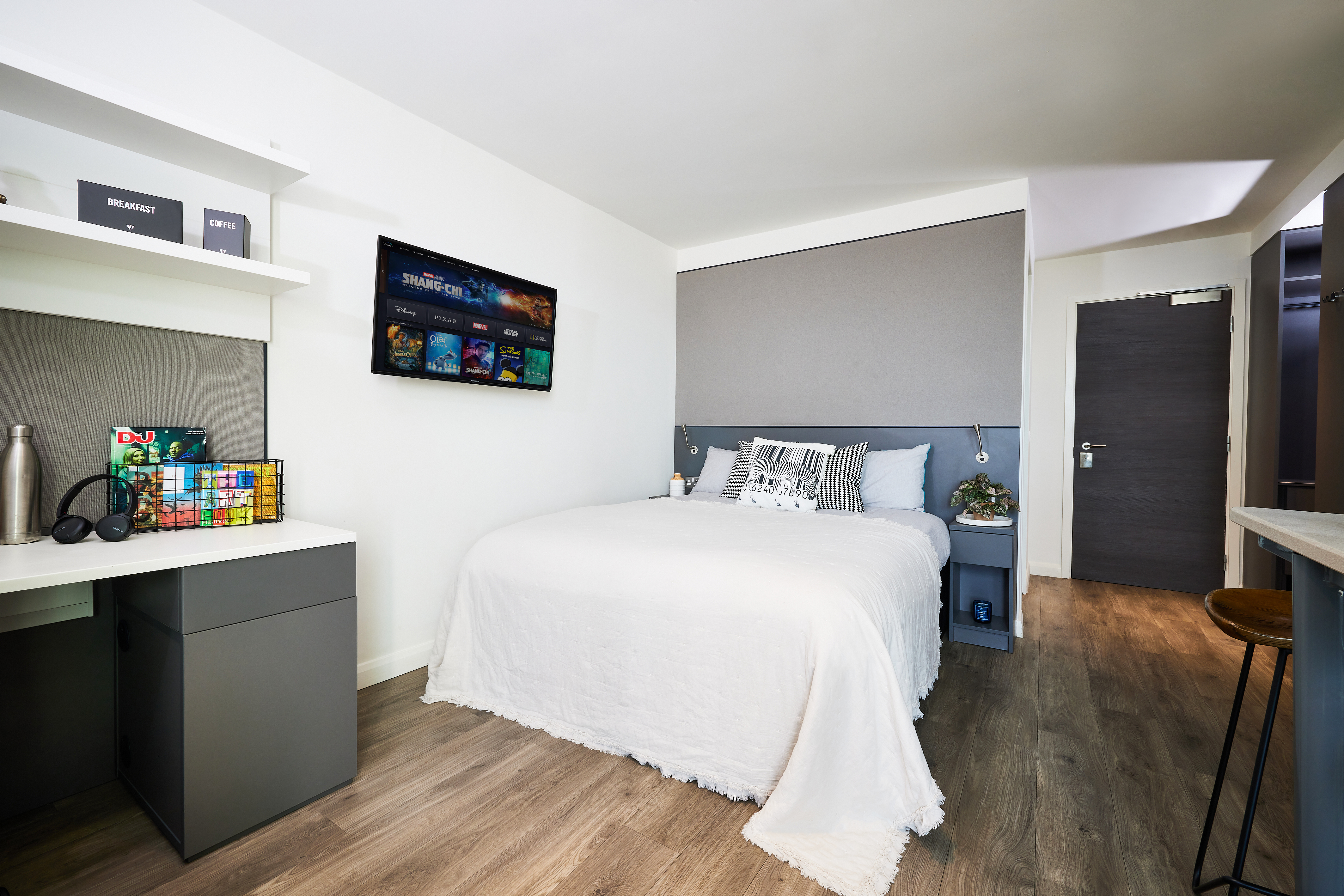 A modern bedroom features a white bed with a gray headboard and wooden floors. A wall-mounted TV showcases movie covers, while a Rubik's Cube and water bottle find their place on the desk. A potted plant and a small bowl of strawberries sit charmingly on the bedside table.