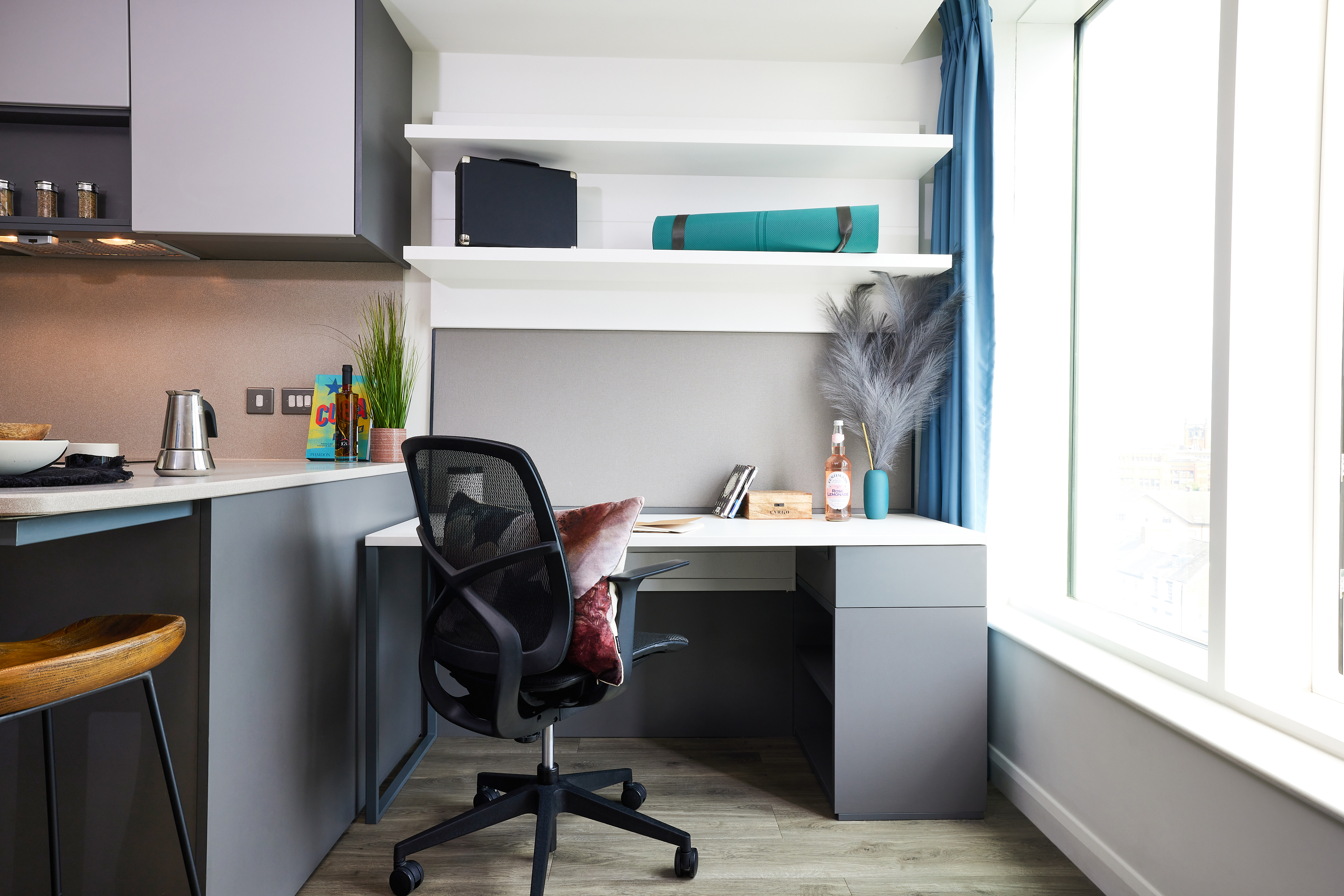A modern home office at Strawberry Place features a gray L-shaped desk with a black office chair, a computer, and a notebook on it. Above are white shelves with decor. Sunlight streams through a large window with blue curtains, illuminating the wooden floor.