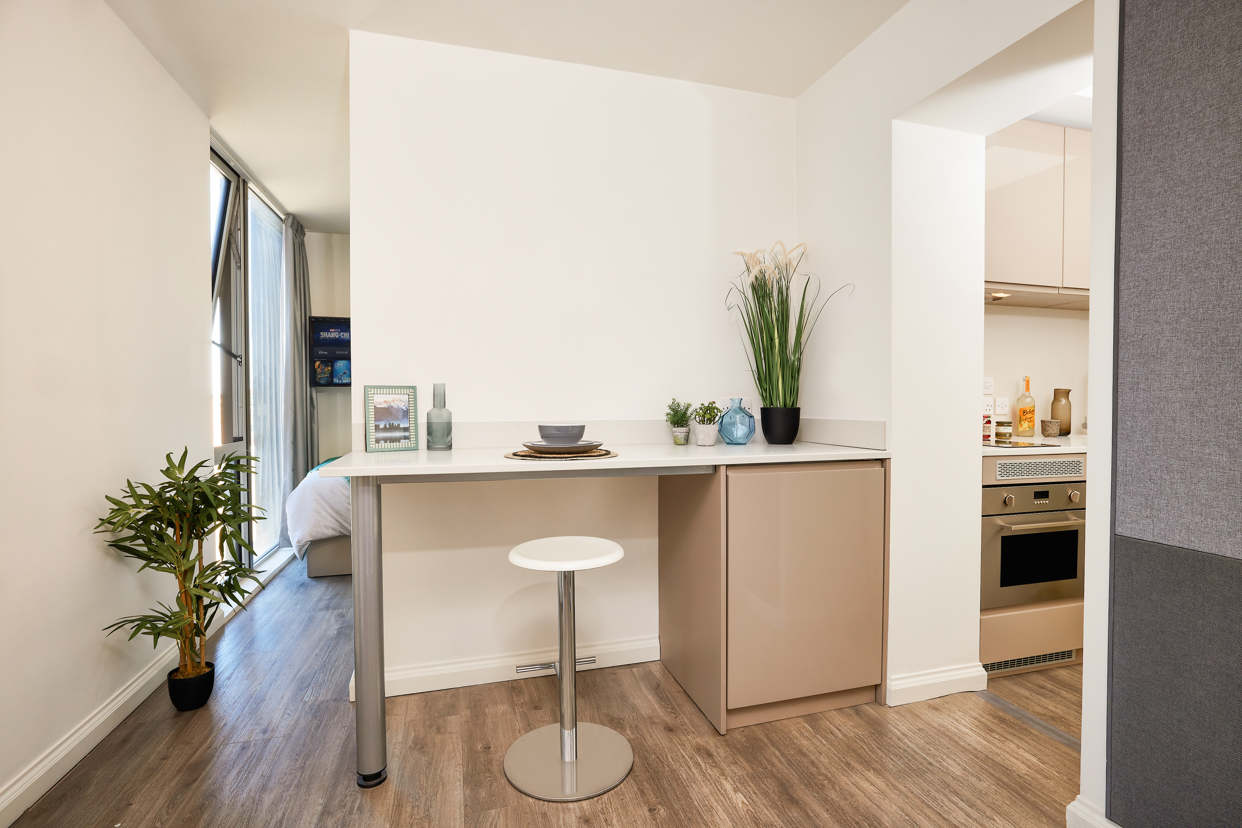 Modern apartment kitchen with a small bar table and stool, reminiscent of an elegant telephone house. The area features potted plants, decorative items, and plates on the table. In the background, a cozy bedroom is partially visible. The space is well-lit with natural light.