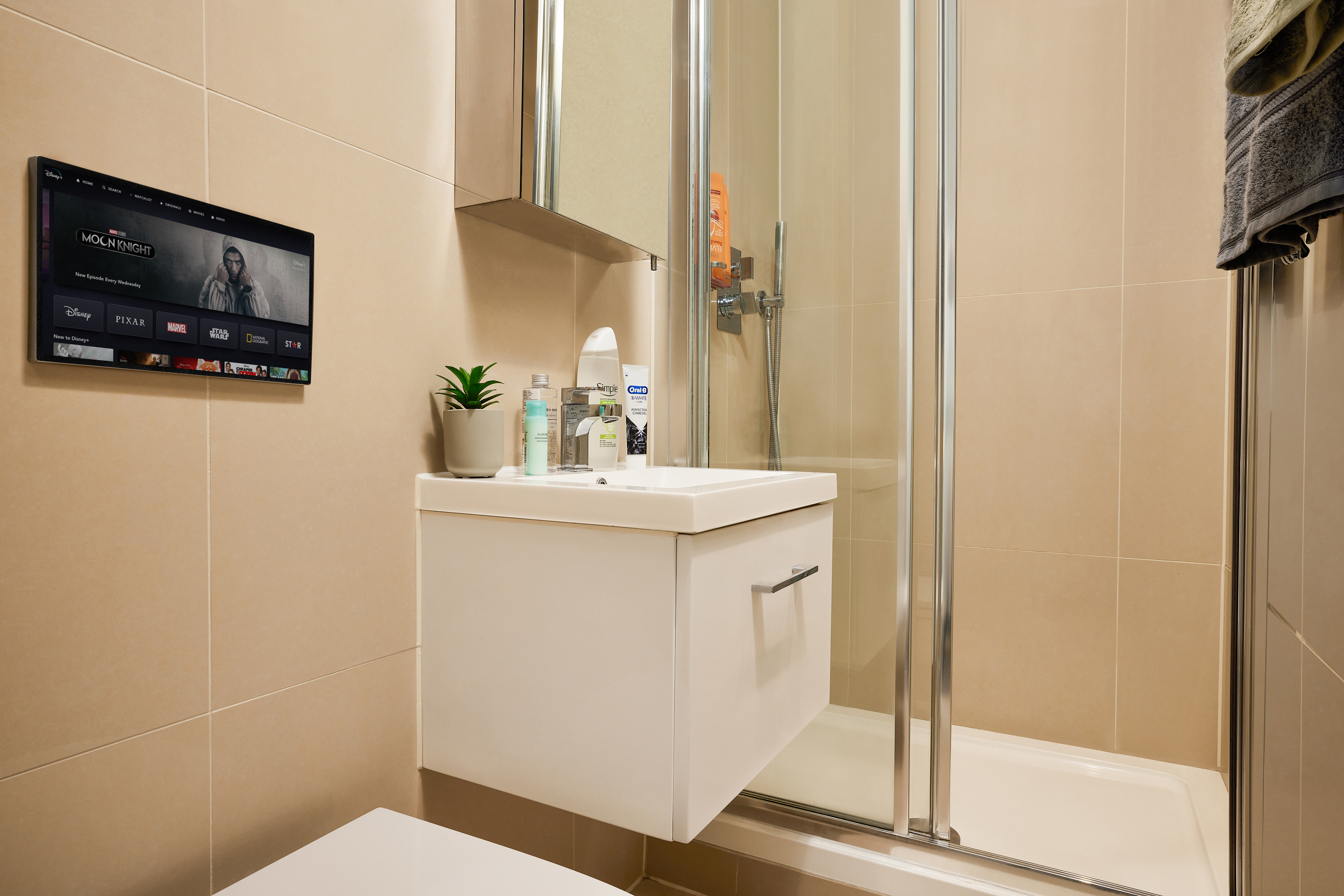 Modern bathroom with beige tiles featuring a wall-mounted screen streaming content from Telephone House. A small, white sink with toiletries, a potted plant, and a mirror above add charm. On the right, a shower with clear glass doors complements the look, while a towel hangs conveniently nearby.