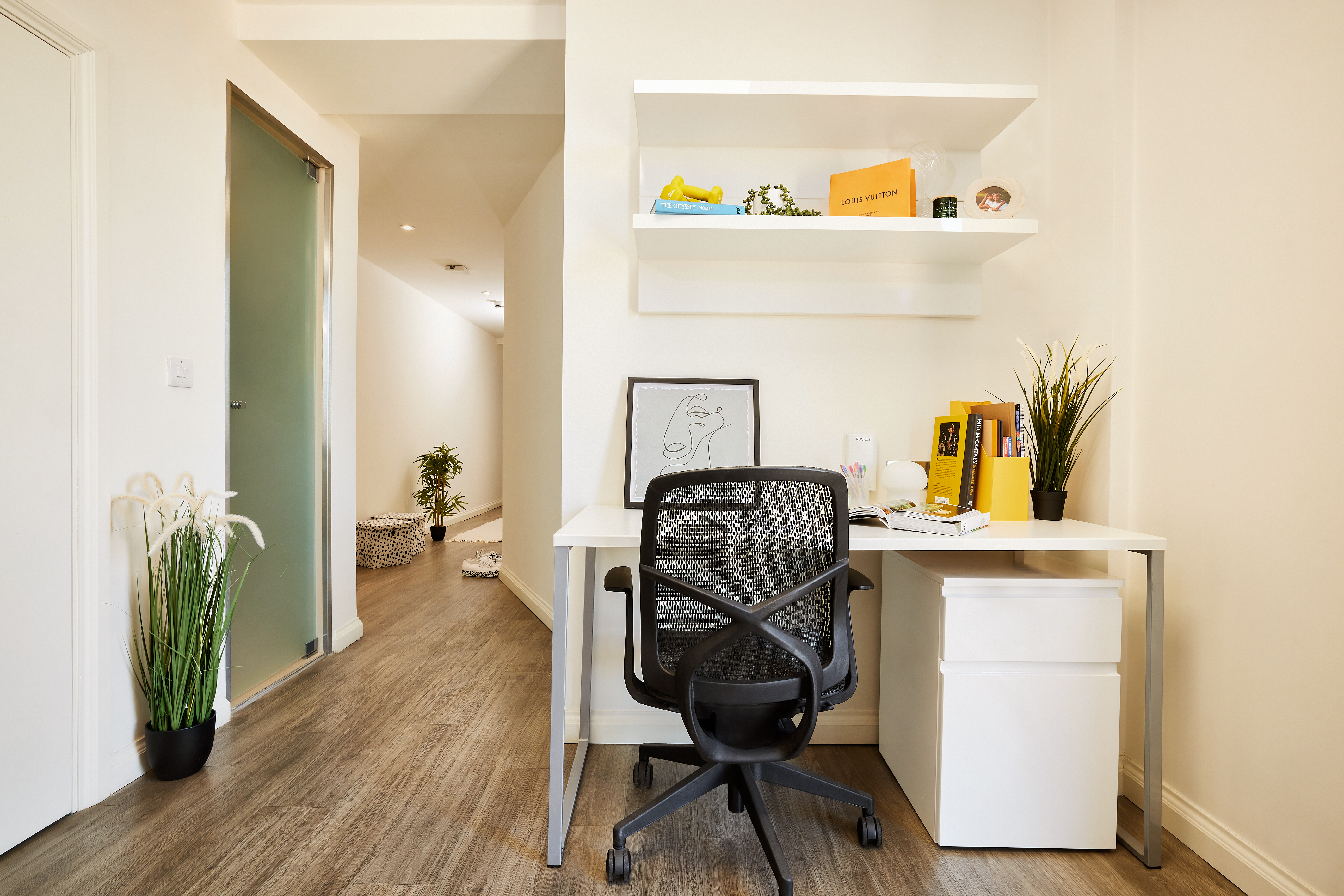 A minimalist home office in Telephone House features a white desk, black swivel chair, and a potted plant. Shelves above display books, a clock, and decor. A hallway with light wood flooring leads to a distant area with a large basket and more plants.