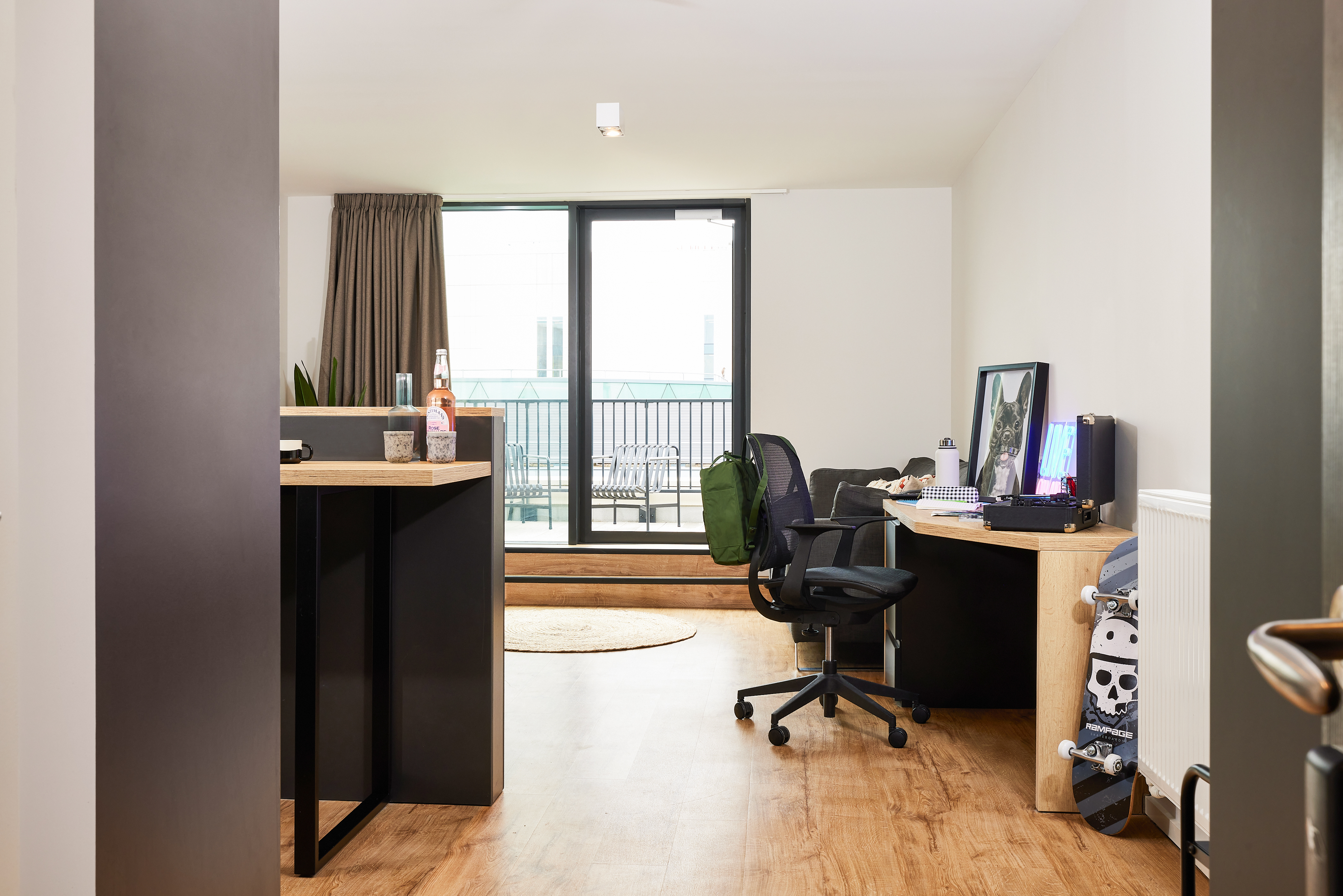 A modern home office in the heart of Portland features a desk equipped with a computer, books, and a backpack on the chair. A skateboard leans against the desk. The room boasts wooden flooring, and a large window opens to a crescent-shaped balcony with chairs and a table.