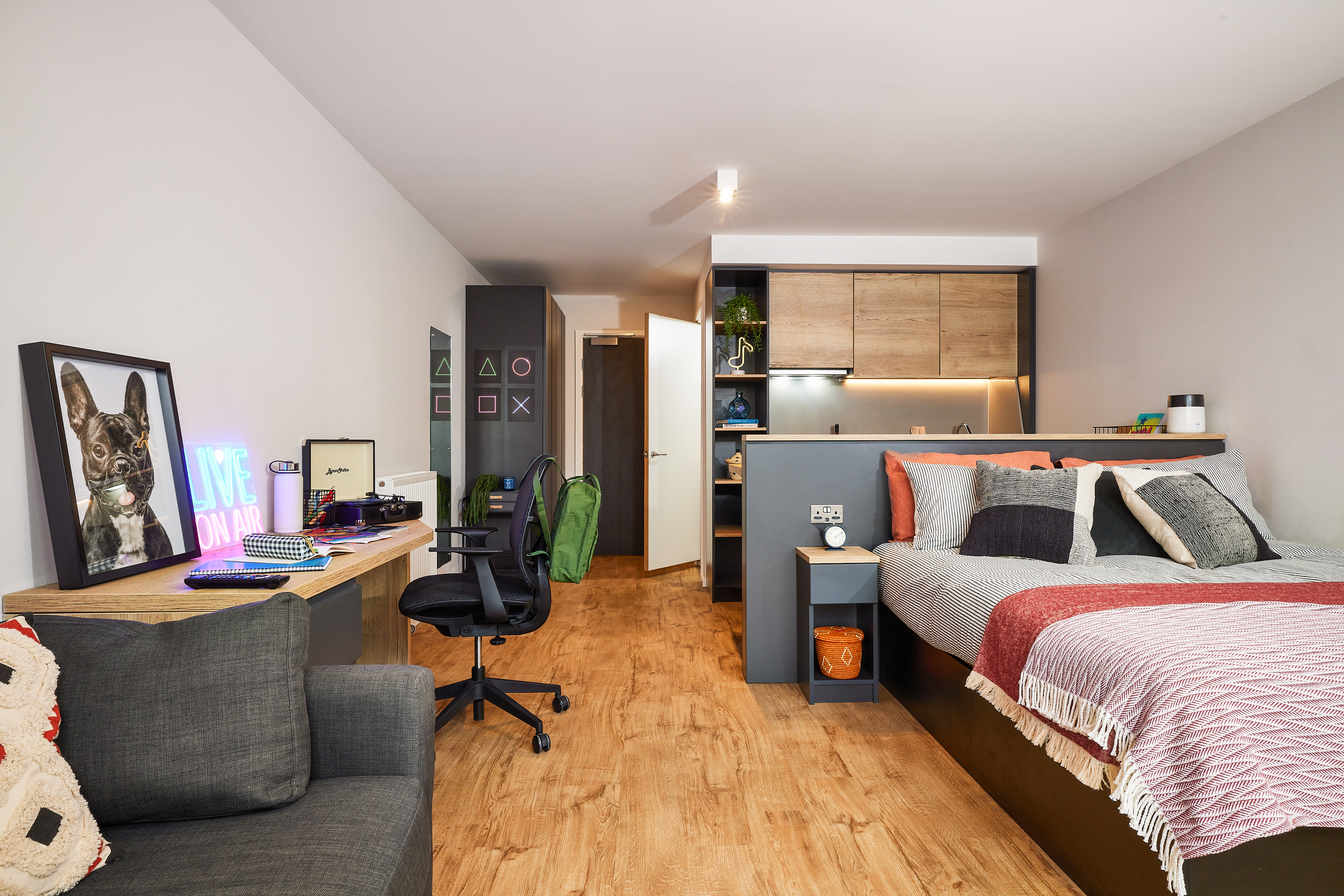 Modern studio apartment in Portland featuring a bed with colorful pillows, a desk with a gaming setup including Crescent neon lights, and a cozy couch. The space boasts wood floors, beige walls, and shelves with plants and decor, creating a warm and organized ambiance.
