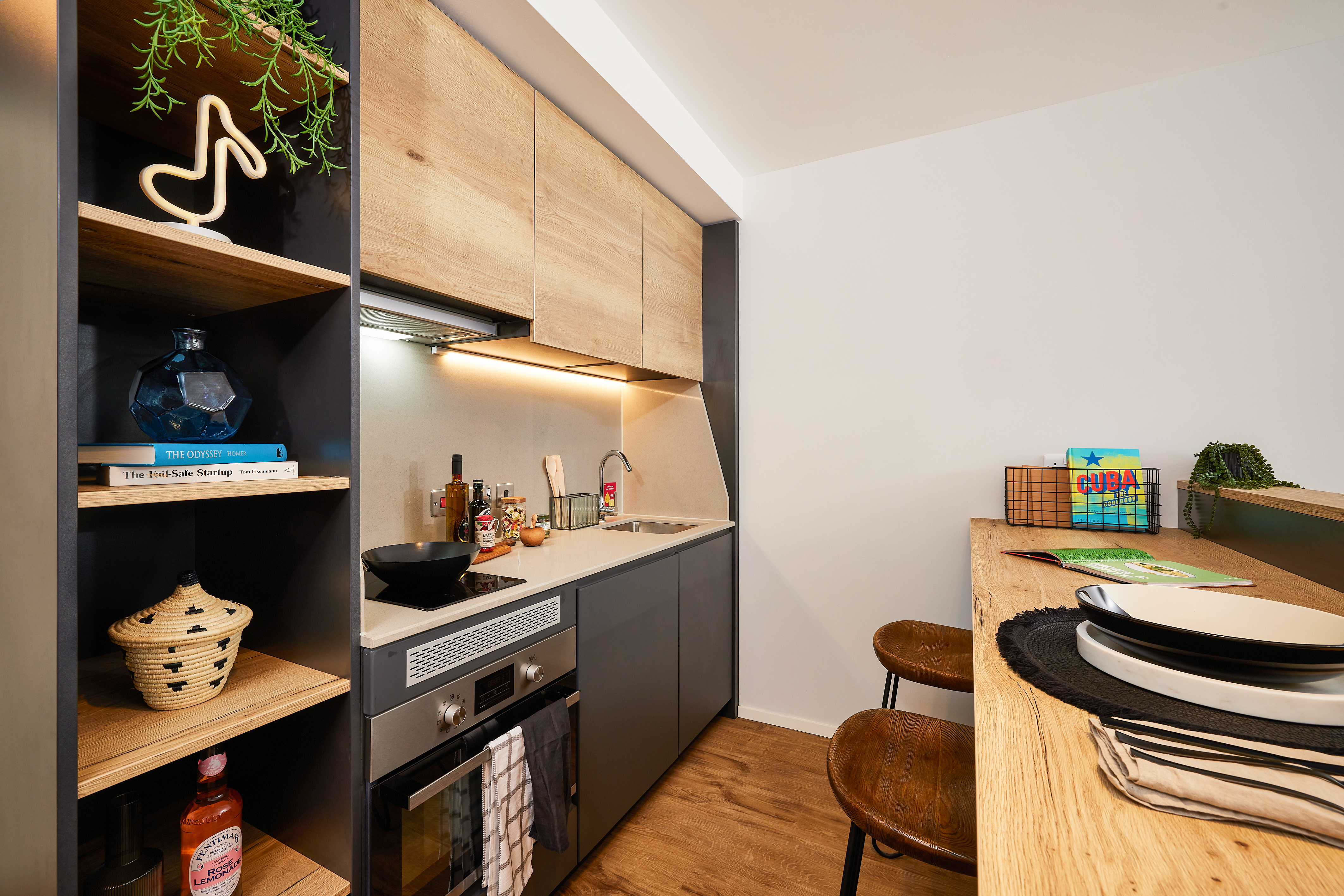 A modern kitchen on Portland Crescent features wooden cabinets and black appliances. Shelving holds cookbooks and decorative items, while a wooden counter with two stools is set with plates and glasses. The space is well-lit and contemporary in style.