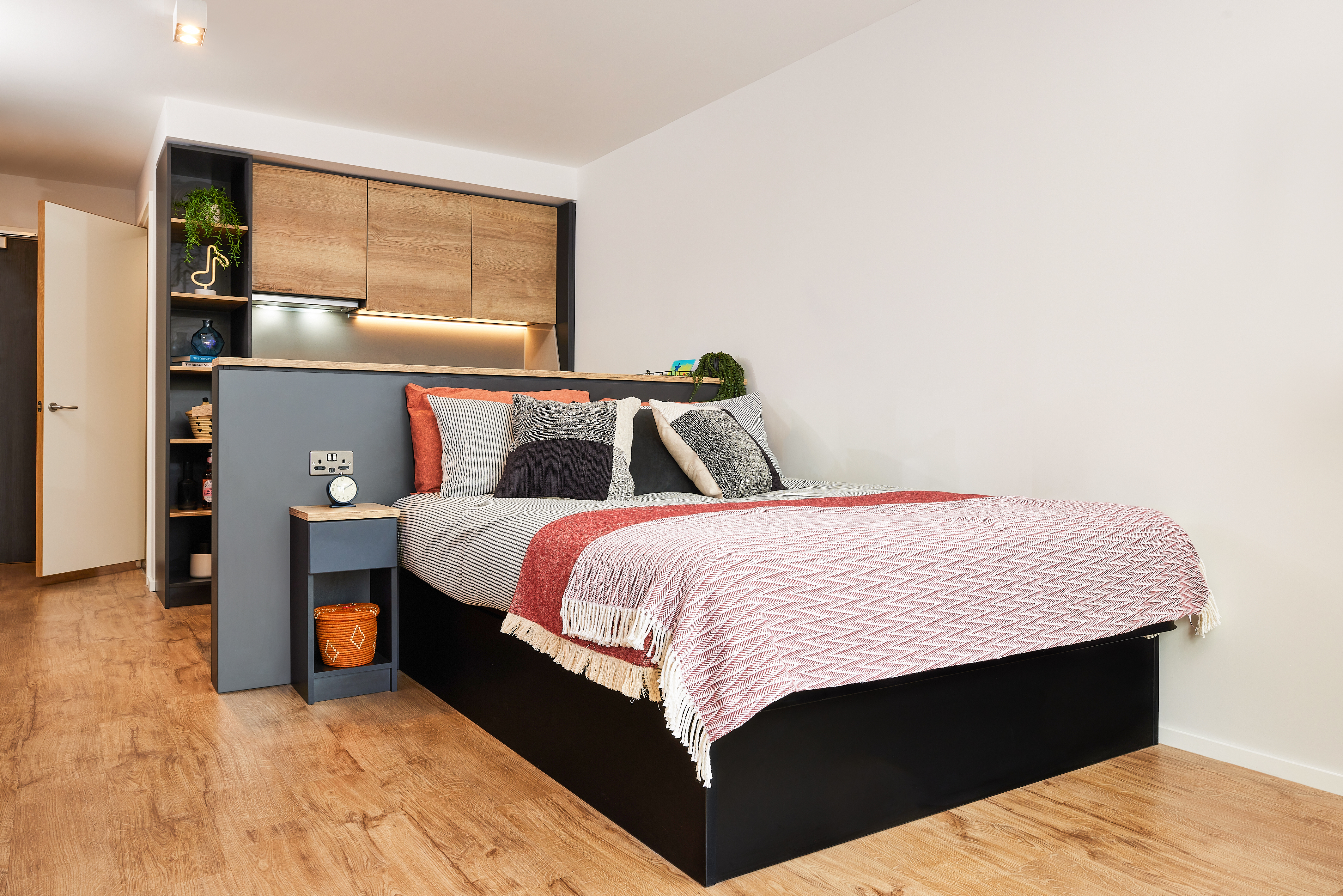A modern bedroom with a wooden floor and a cozy bed, draped in a red and white patterned blanket, offers comfort. A crescent moon-shaped bedside table holds a clock and decor items. Open shelving and soft lighting add warmth to this Portland-inspired space.