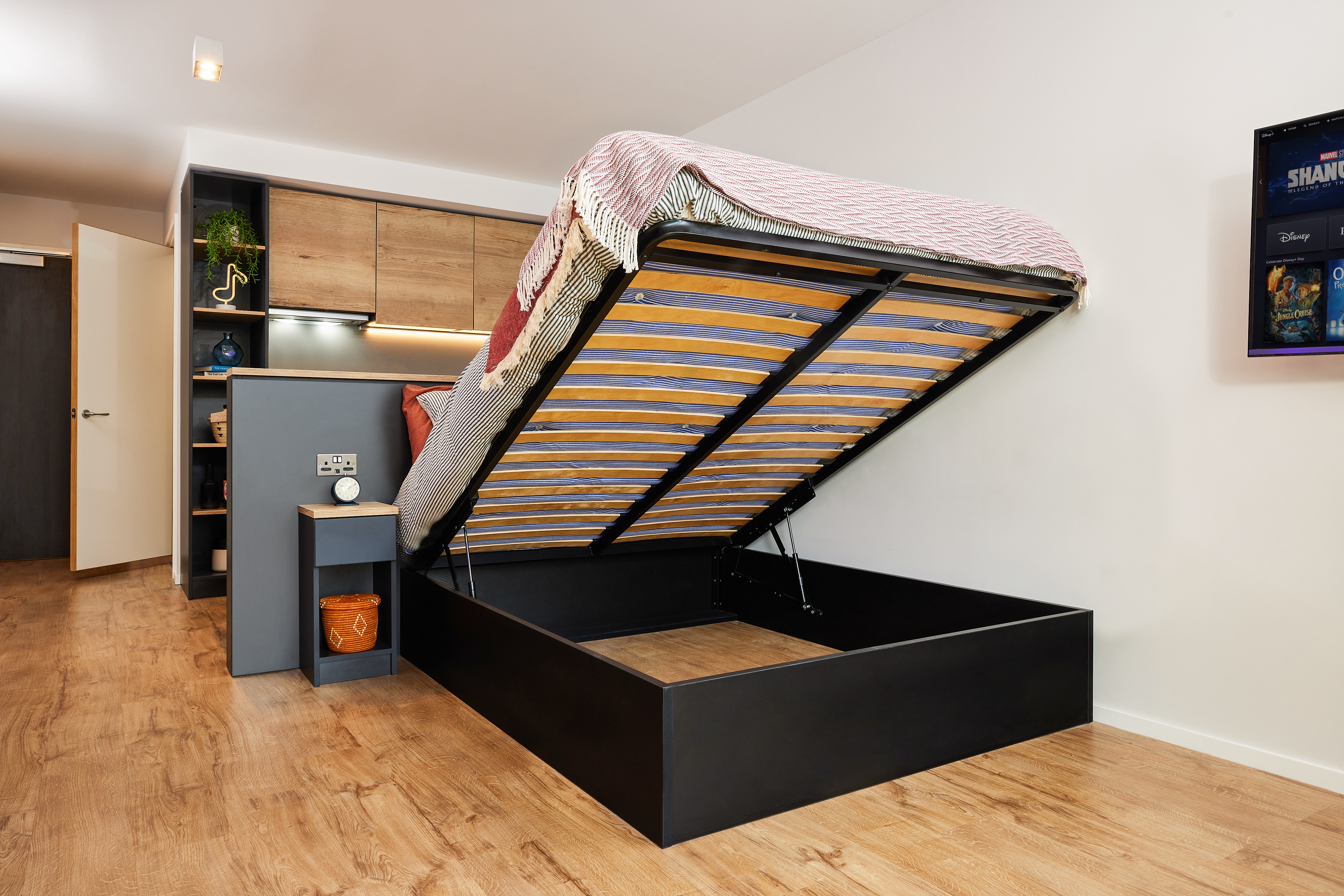 A modern bedroom in Portland showcases an open lift-up storage bed, revealing ample space. The bed has wooden slats and a black frame. The room includes wooden flooring, built-in furniture, and a wall-mounted TV, creating a cozy crescent of comfort.