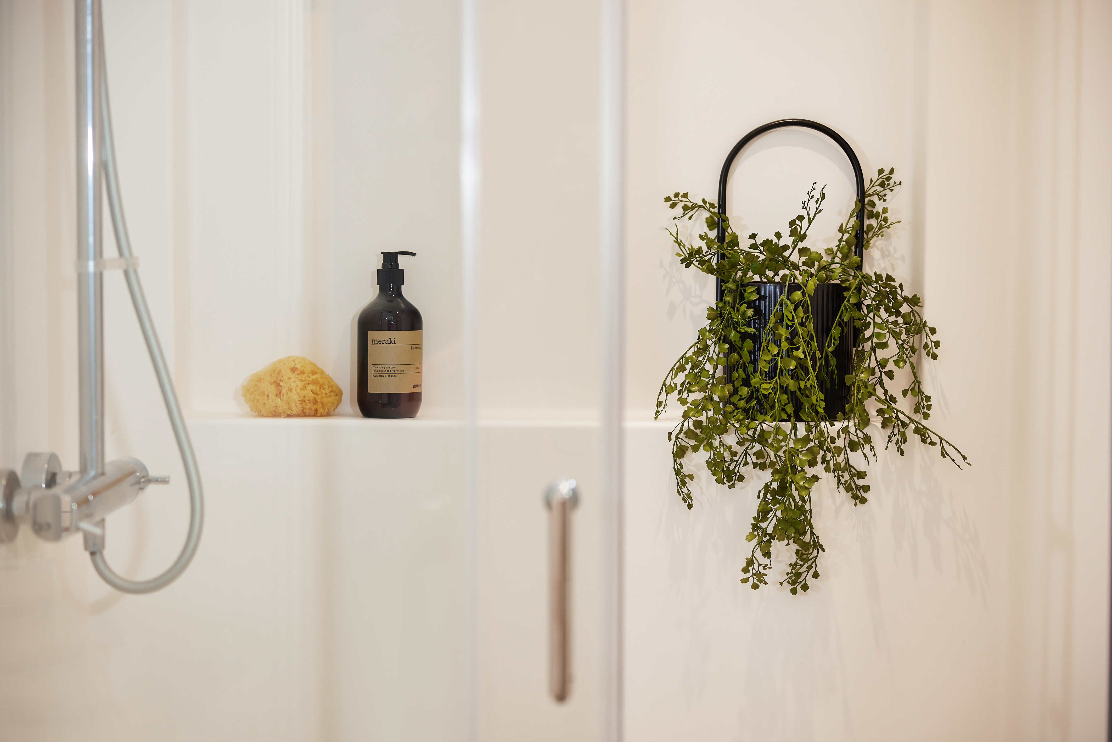 In the Portland House style, this minimalist shower stall features a clear door. Inside, a black bottle of soap and a natural sponge rest on the ledge. On the wall, a black vase with trailing green foliage adds a touch of nature to the space.