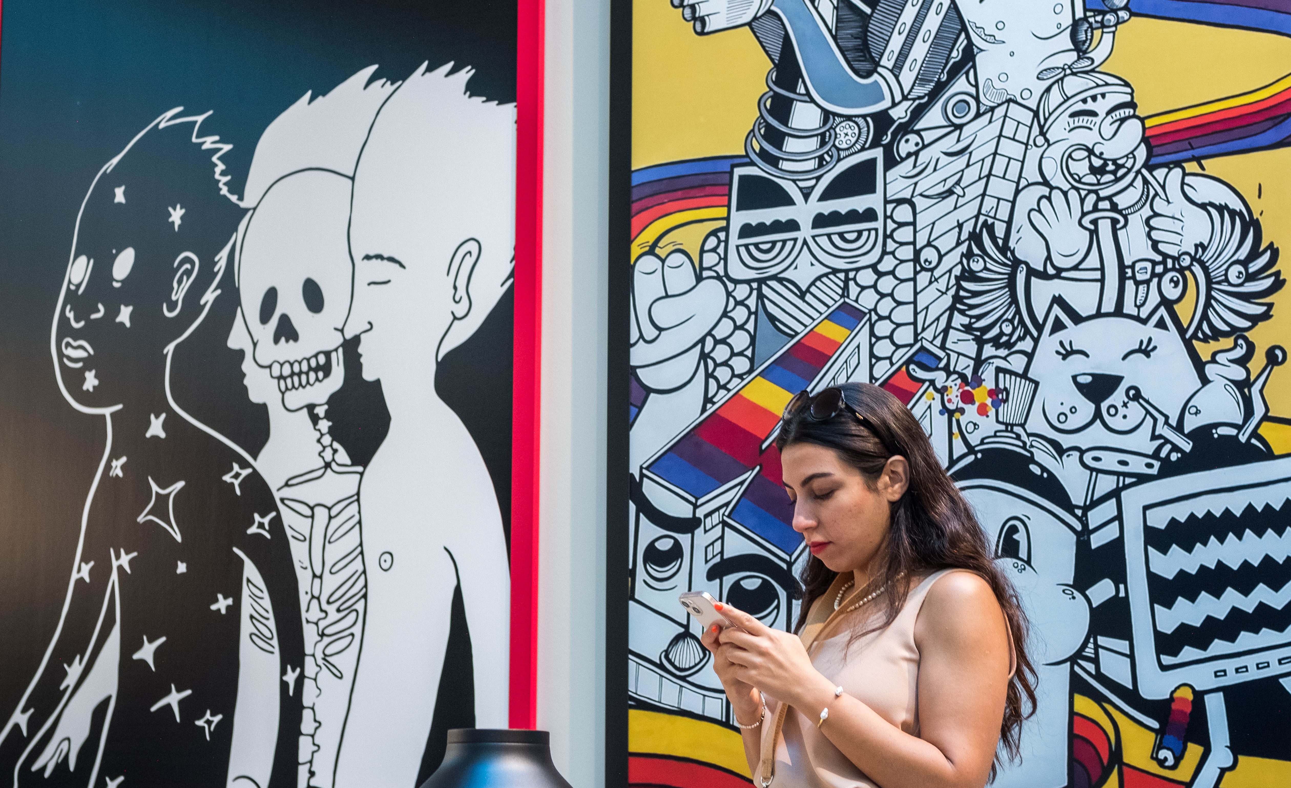 A woman looks at her phone while standing in front of colorful street art. The mural features cartoonish characters, including a skeleton, a robot, and a cat against vibrant backgrounds. She wears sunglasses on her head and is holding earphones.