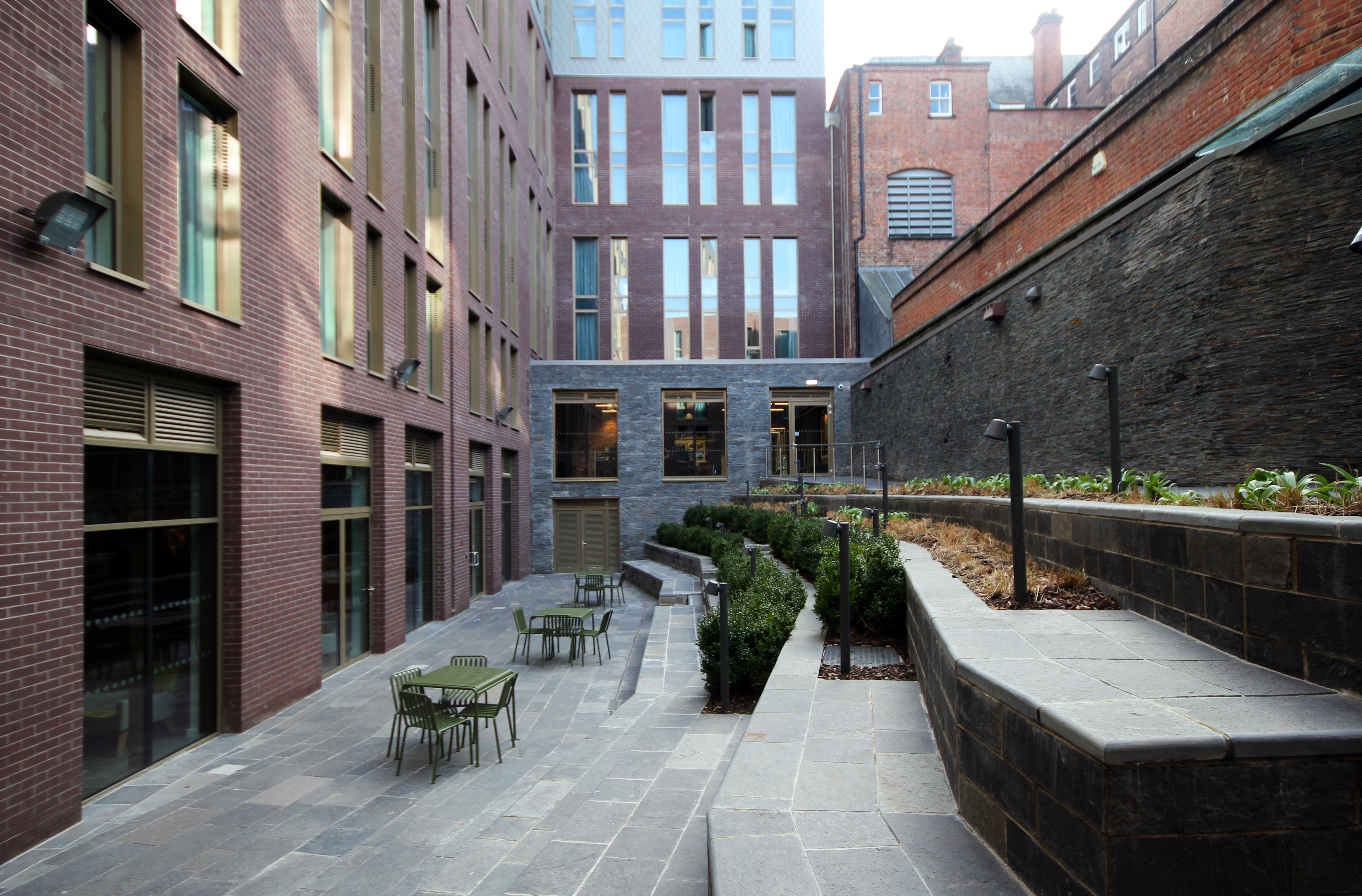 A modern urban courtyard with tall brick buildings on either side. The space features stone pathways, outdoor seating with green tables and chairs, and planters with small shrubs. Large windows reflect a clear sky above.