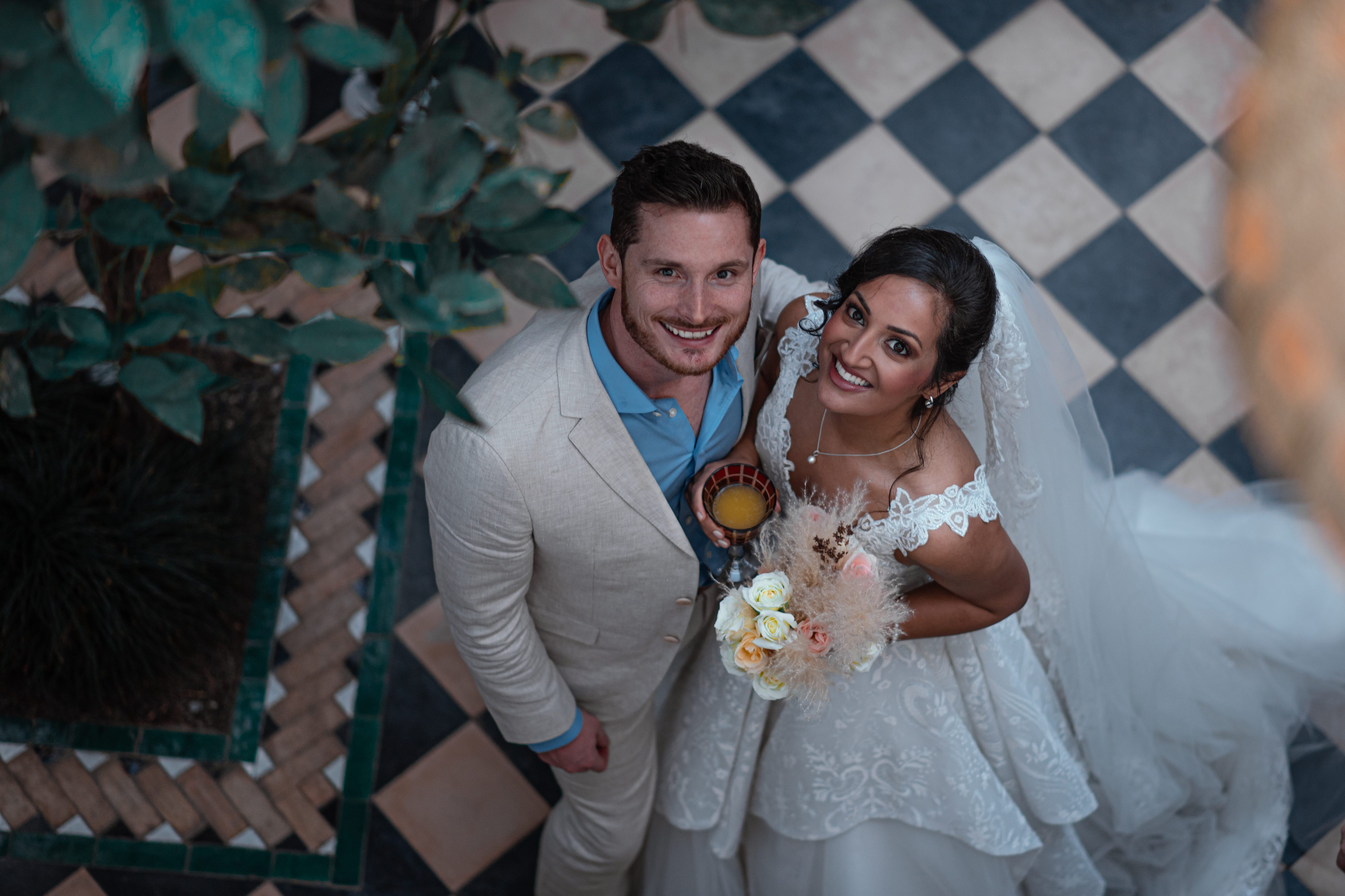 A couple stands smiling, looking up at the camera. The man, having just married in Marrakech, wears a light beige suit while the woman is in a white wedding dress, holding a bouquet. The floor beneath them showcases a charming checkered pattern reminiscent of Vita Student’s creative spaces.