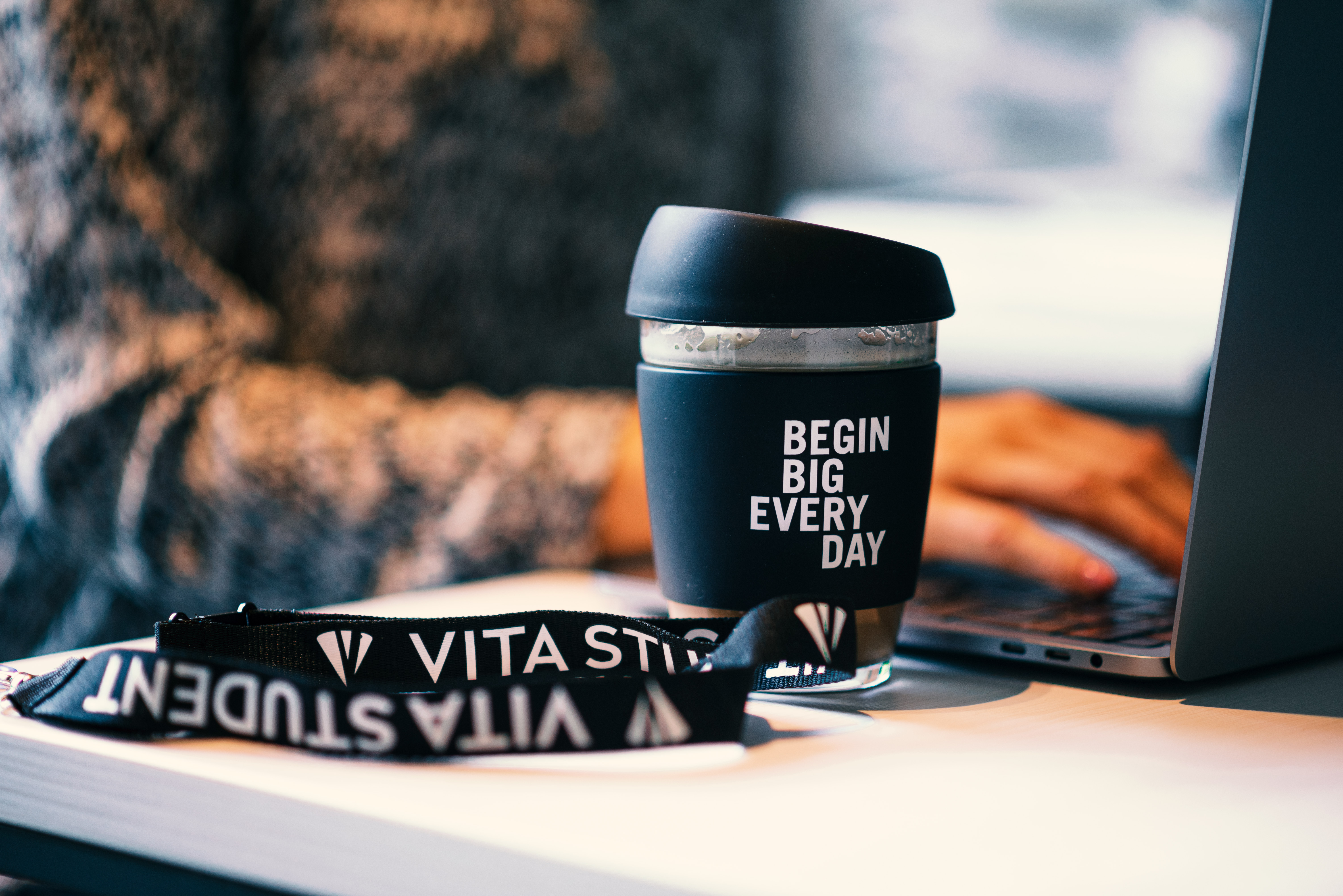 A person working on a laptop with a reusable coffee cup that reads "BEGIN BIG EVERY DAY" on the desk. A lanyard with "VITA STUDI" is also on the table.