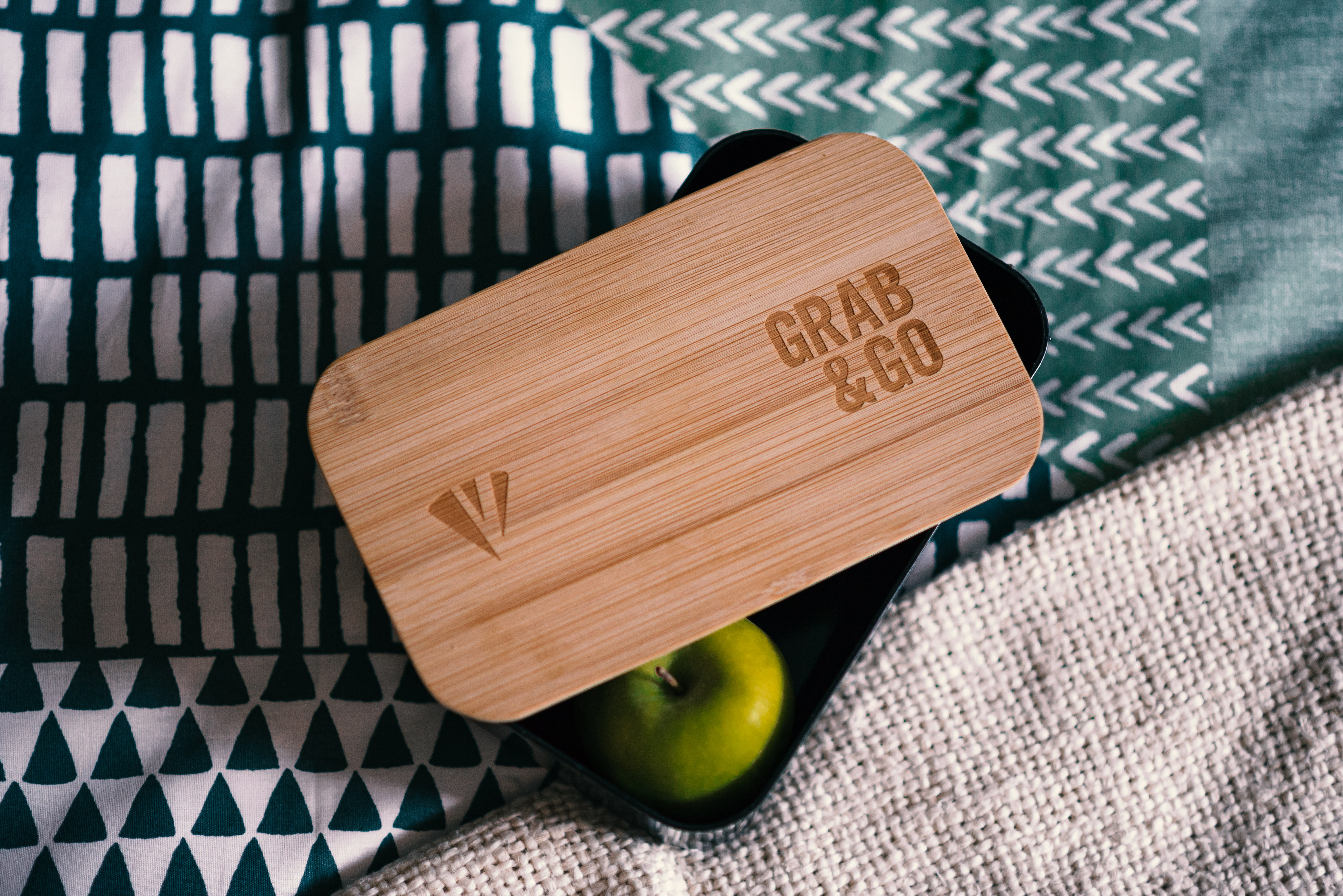 A rectangular wooden lunchbox lid with "Grab & Go" engraved on it rests on a patterned fabric. The lid is slightly open, revealing a green apple inside the black lunchbox. Designed in EN-UK style, the fabric boasts geometric patterns in teal and white.