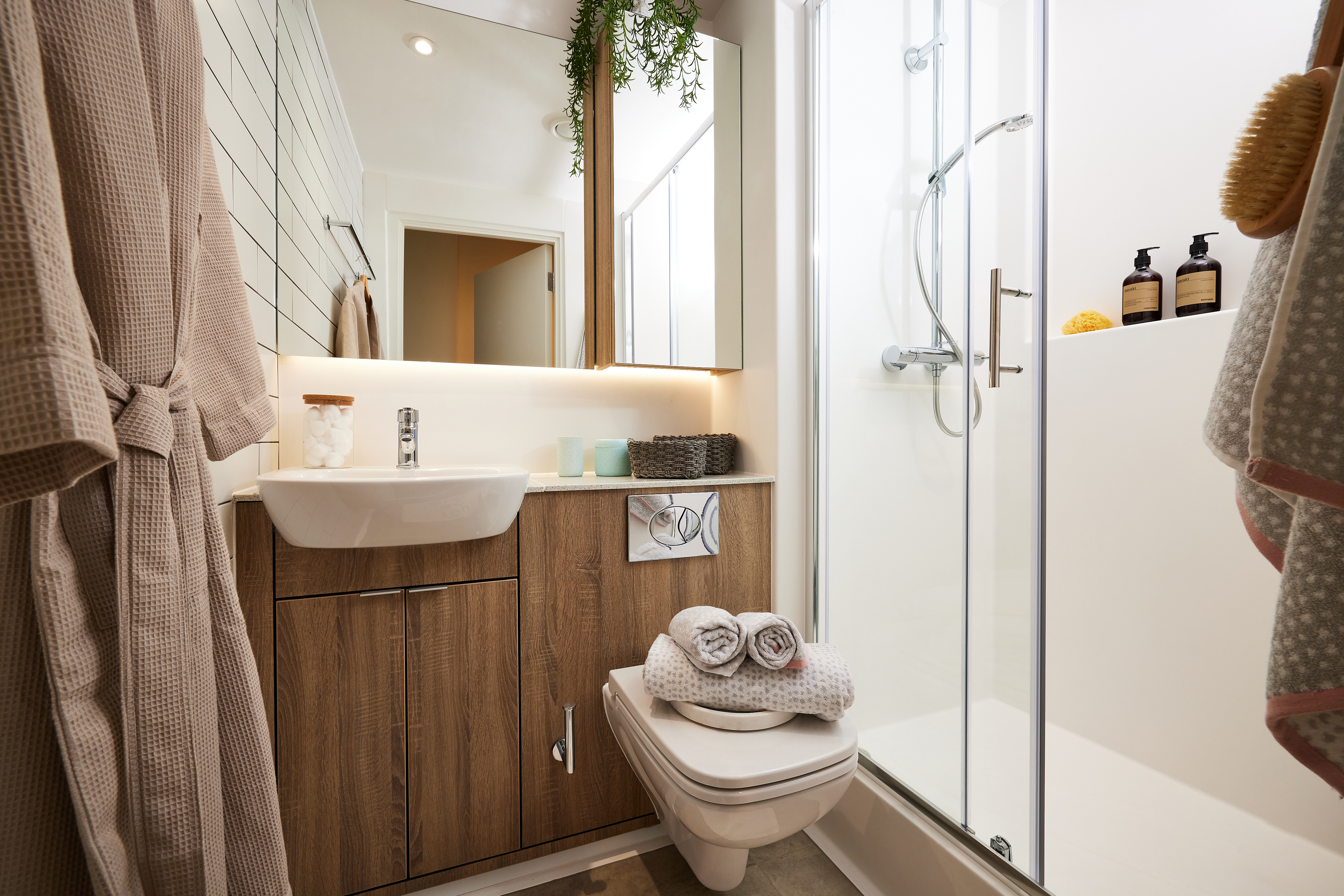 A modern bathroom features a Warwick wood-paneled vanity, white sink, and towels. A wall-mounted toilet and glass-enclosed shower are on the right, with robes hanging on the left. Lush greenery adorns above the mirror, while Cannon Park-inspired bottles grace the shower.