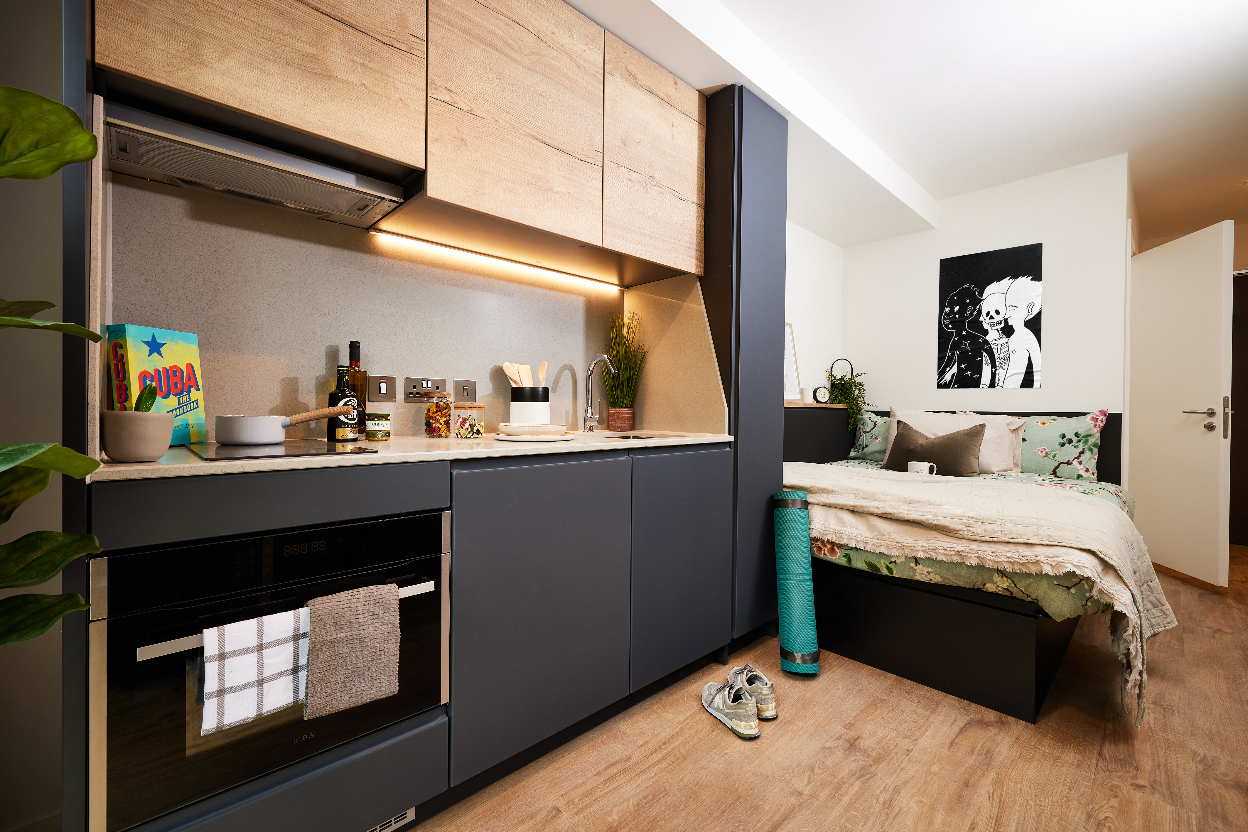 A compact studio apartment near Warwick features a small kitchen with wooden cabinets, a cooktop, oven, and countertop appliances. Adjacent is a cozy bed with floral bedding, a rolled yoga mat, and a potted plant. Monochrome wall art completes this charming Cannon Park retreat.