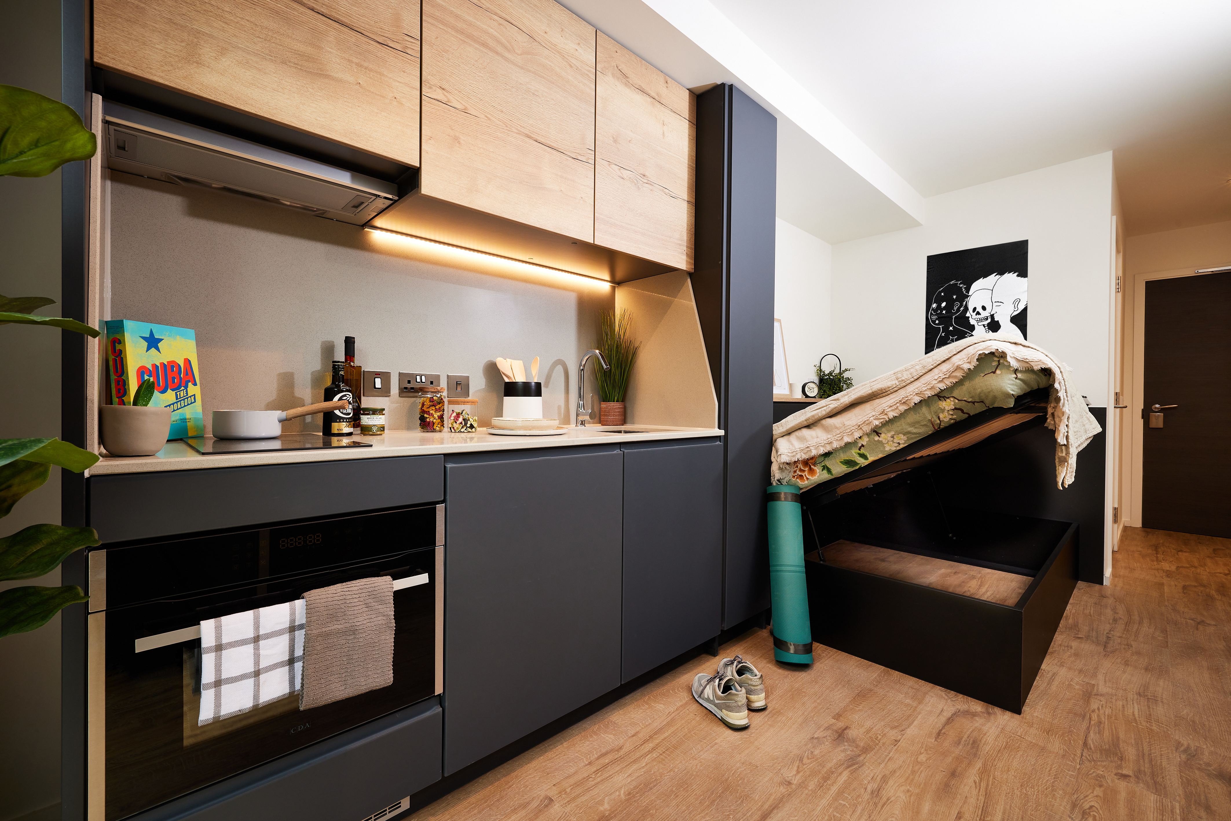 Modern studio apartment in Copper Towers with a compact kitchen featuring wooden cabinets, a black oven, and countertop appliances. A bed with storage underneath is partially lifted. A green yoga mat and sneakers are on the wooden floor. Wall art and a potted plant add decor.