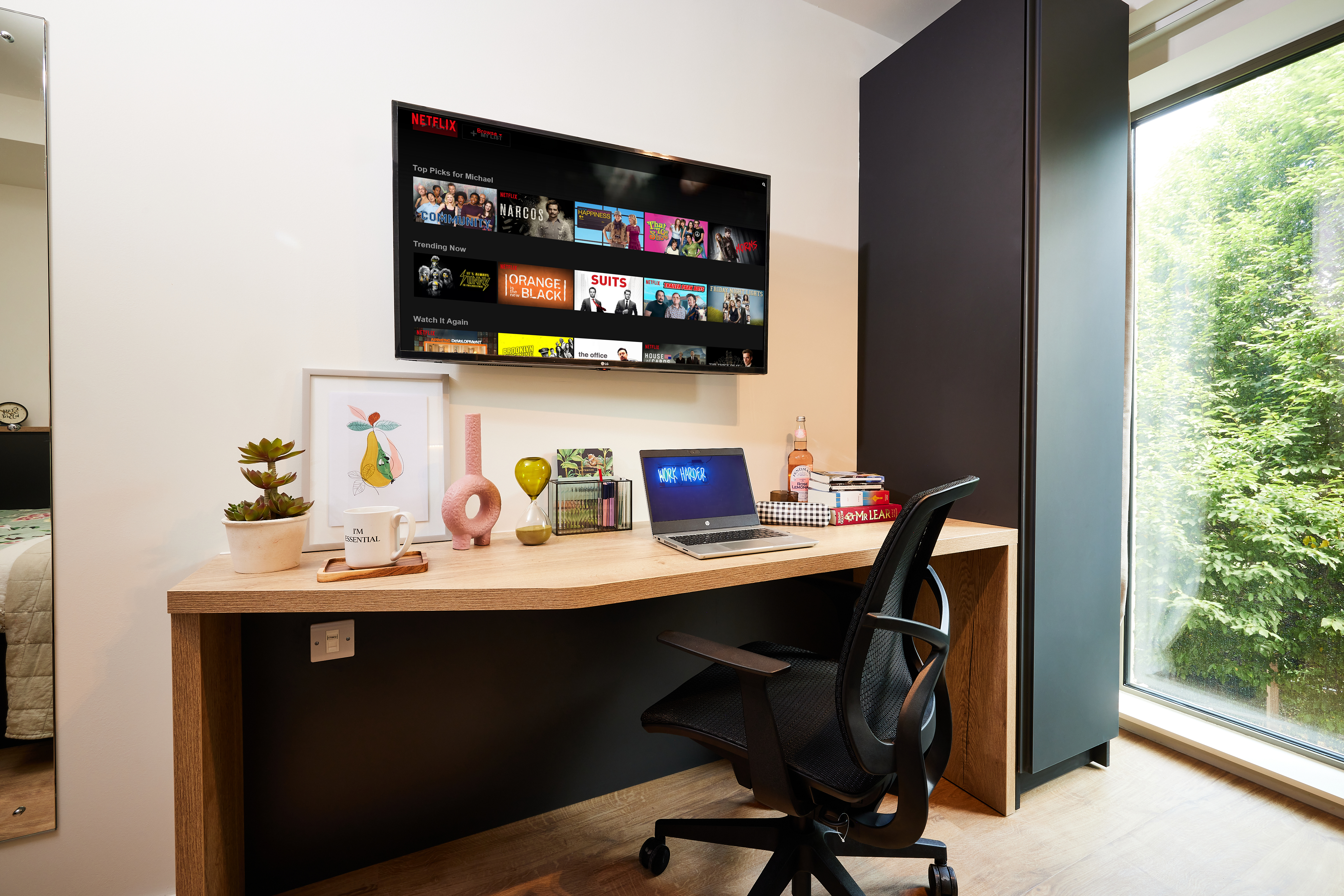 A modern home office in Cannon Park features a wooden desk, black chair, and laptop. Plants and decorative items adorn the desk. A TV displaying a streaming service interface is mounted on the wall, while a large window showcases Warwick's greenery, flooding the space with natural light.