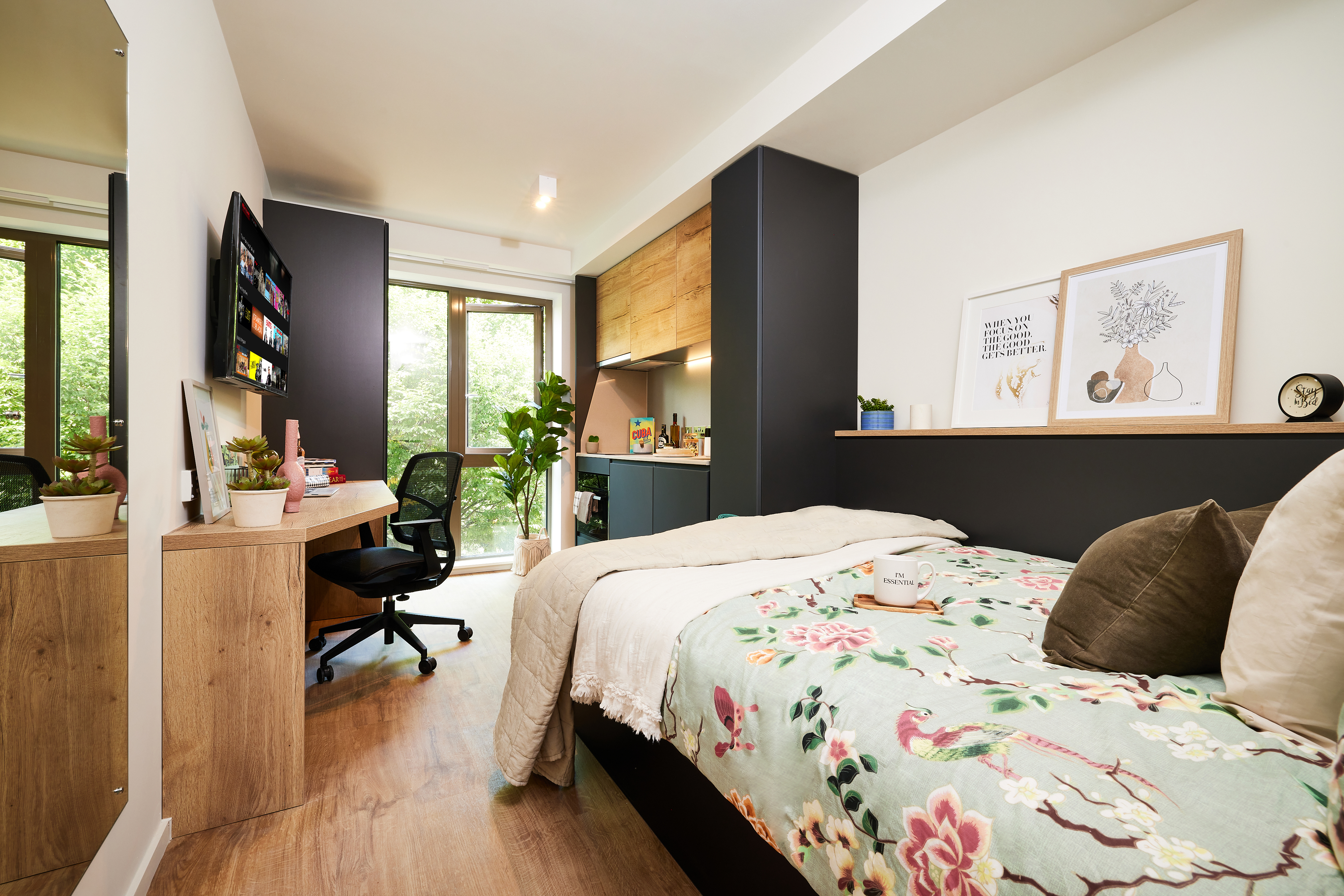 In a modern bedroom near Cannon Park, with a neatly made bed adorned with pillows and a floral blanket, you'll find a desk, chair, potted plant, and wall-mounted TV beside the window. Art prints and decor items grace the shelf above the bed, all set on light wood flooring throughout.