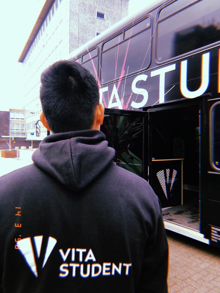 A person wearing a "Vita Student" hoodie stands in front of a black bus with partial text "STUD" visible. The bus door is open, inviting agents to board, while a modern building partners in the backdrop.