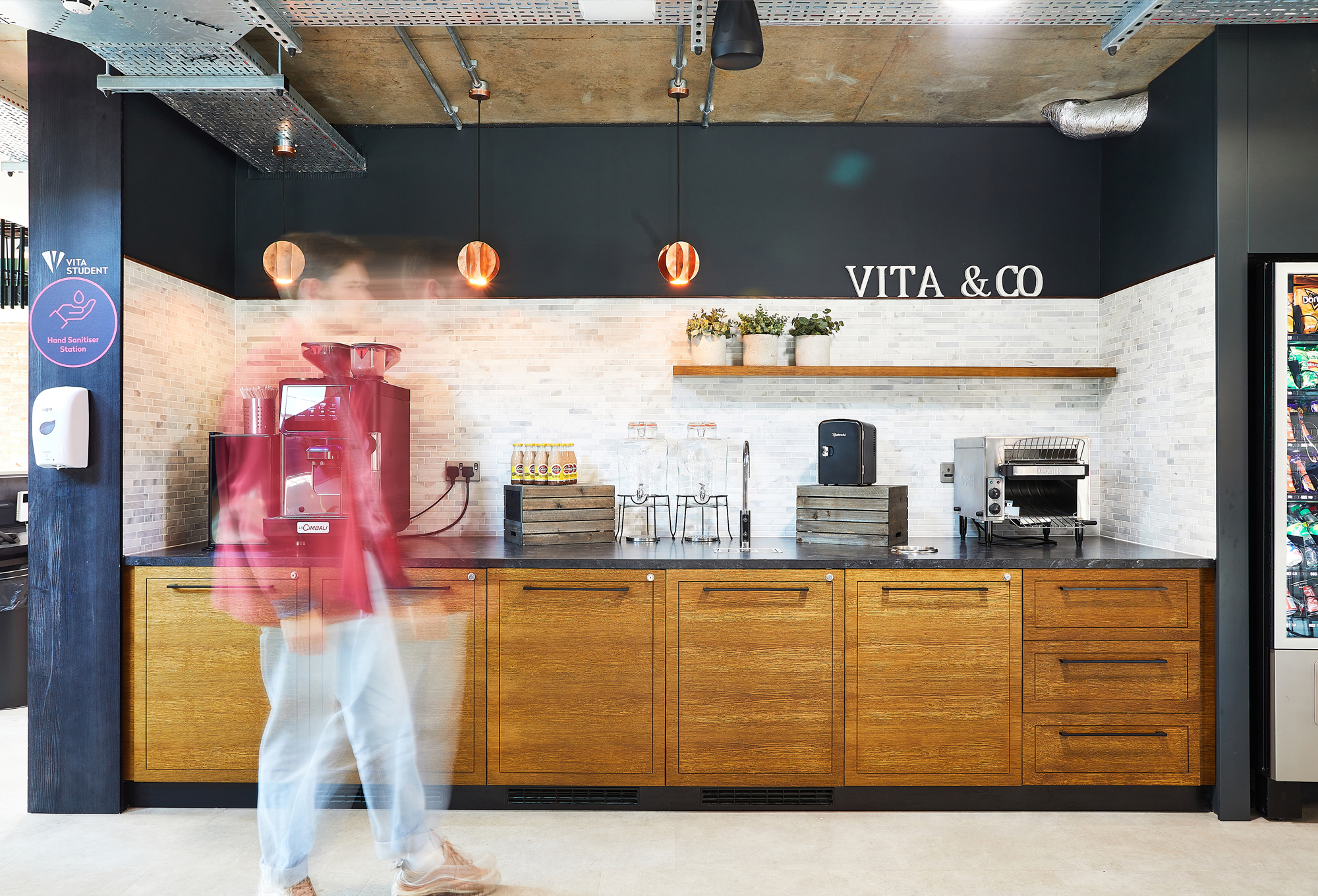 A blurred person strolls through a modern UK kitchen featuring wooden cabinets and a marble backsplash. On the counter, a coffee machine and sanitizer sit beneath a "VITA & CO" sign. Lush plants grace the wall shelf, while bright lighting highlights the industrial decor elements.
