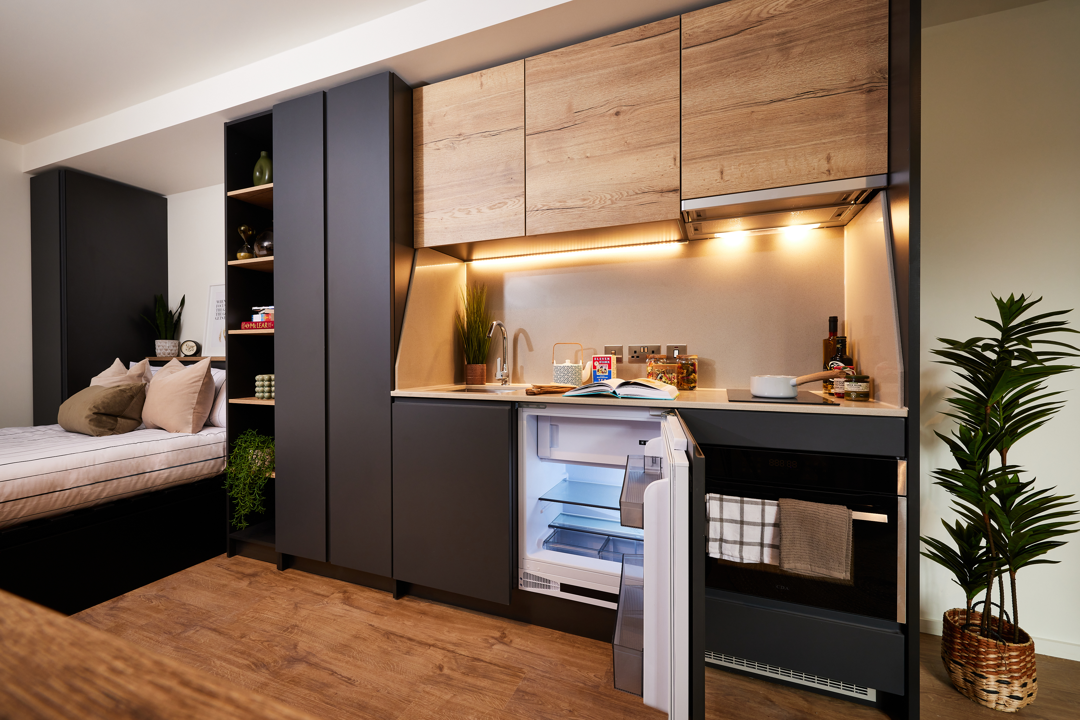Modern studio apartment near Cannon Park with a compact kitchen featuring dark cabinets, wood accents, and under-cabinet lighting. Open fridge and oven. Cozy bed area with pillows and plants. Hardwood floor complements the potted plant on the right, offering a touch of Warwick charm.