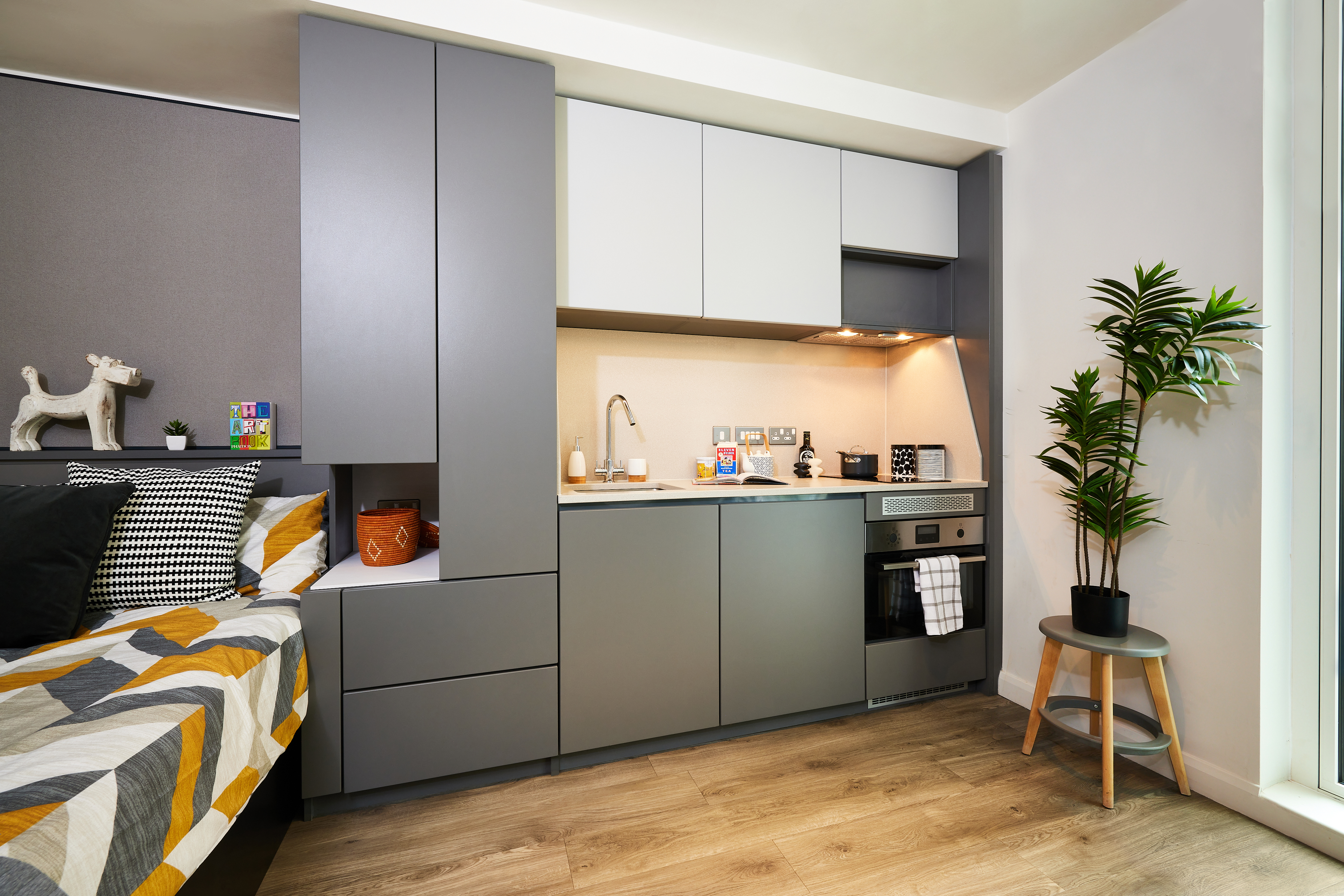 A modern studio apartment features a compact kitchen with gray cabinets, a stove, and a sink. A bed with patterned bedding is nearby, and a decorative plant sits on a small table. The wooden floor adds warmth to the place, complementing the cozy vibe of this urban strawberry haven.