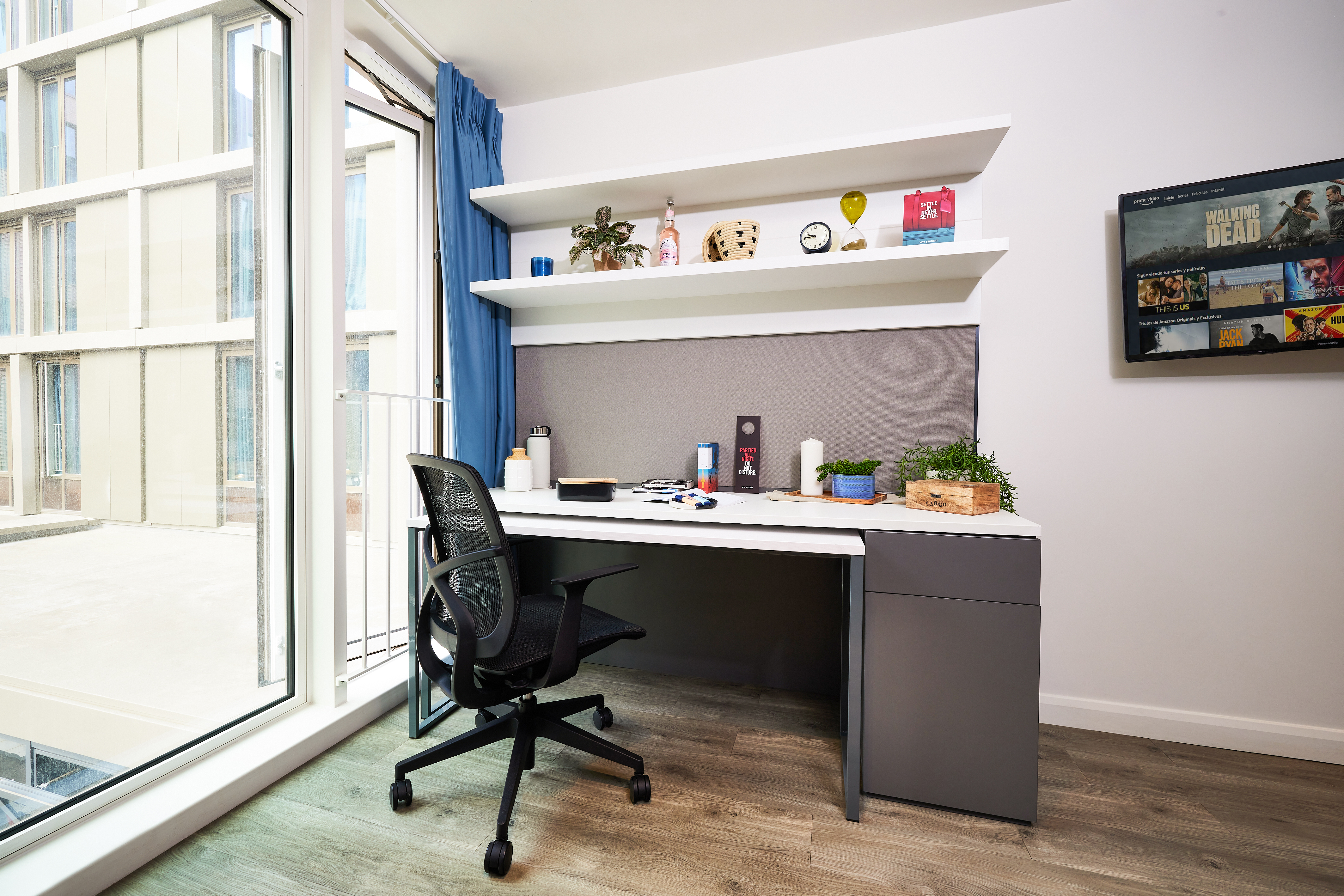 A modern study room features a desk and a black office chair. The desk is adorned with books, a laptop, a lamp, and assorted items. Shelves above boast plants and strawberry decor. A large window offers views of surrounding buildings, while a framed poster graces the right wall.