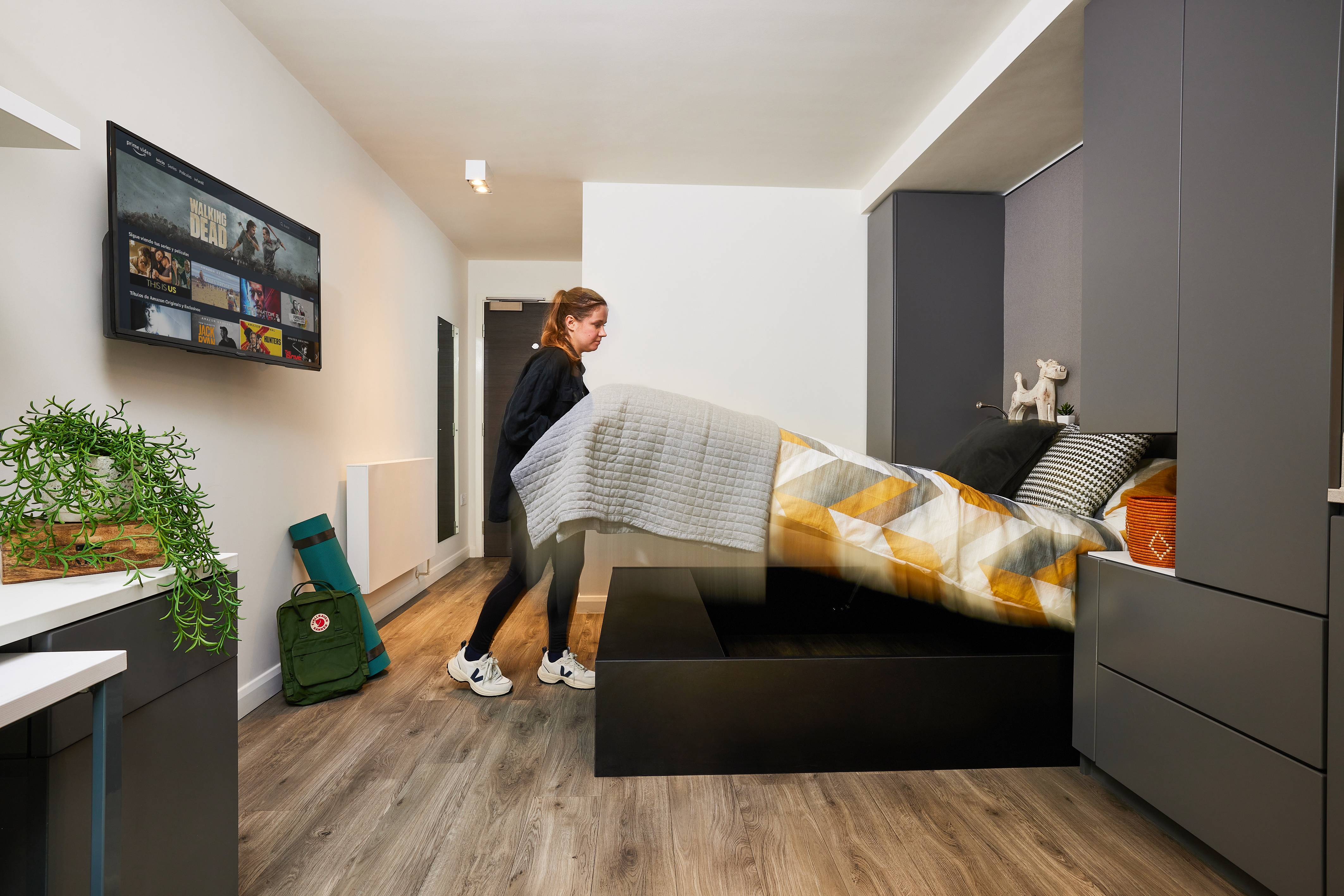 In a stylish room at Strawberry Place, a person is folding a bed on the light wood floor. A TV on the wall displays shows while nearby, a plant and backpack add charm. The decor features gray wardrobes, a shelf with toys, and a neatly made bed with a patterned duvet.