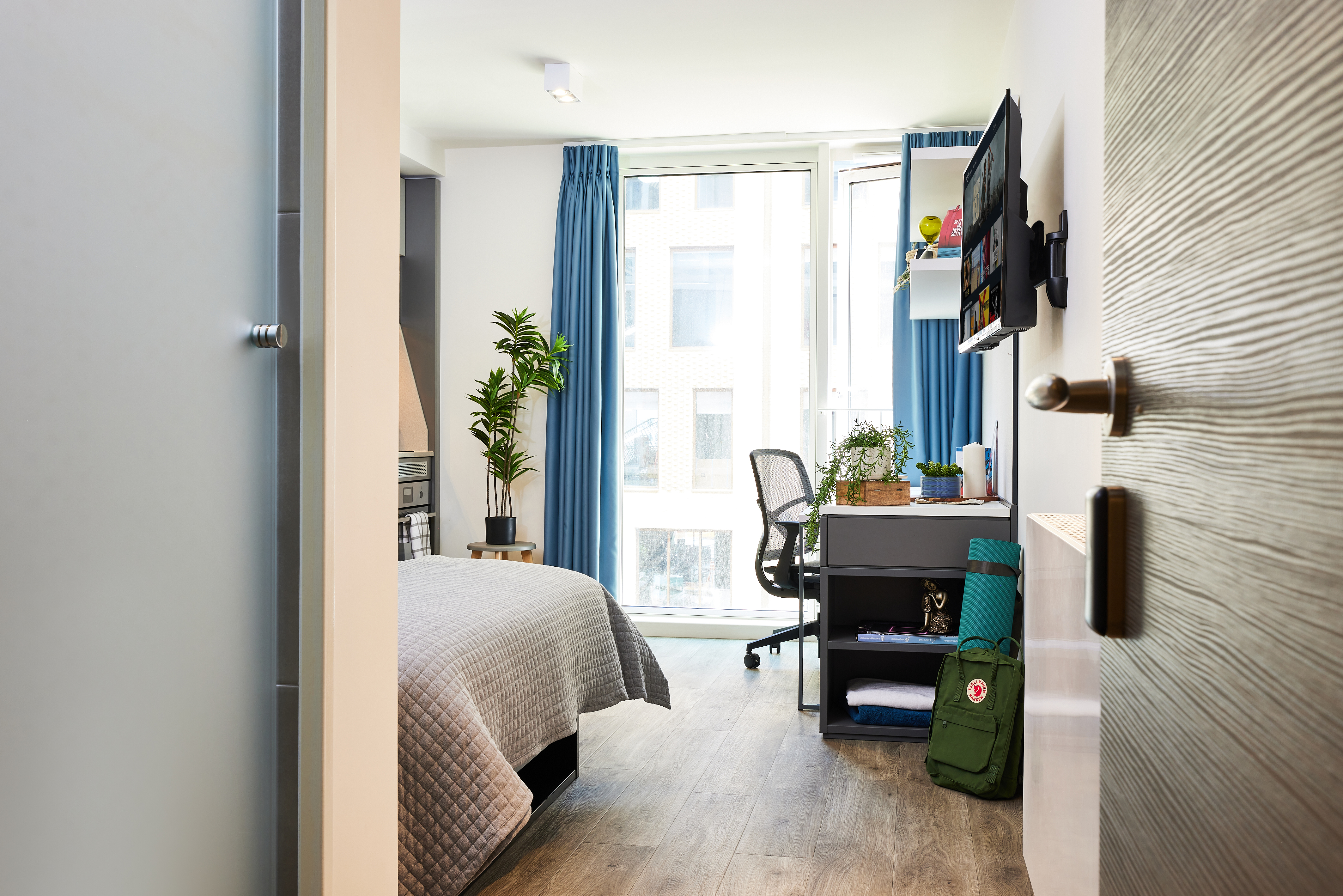 A tidy bedroom in Strawberry Place features a bed, desk, and chair. A TV is mounted on the wall above the desk. Sunlight pours through large windows with blue curtains. A backpack rests on the wooden floor, while plants add a touch of nature to the room's charm.