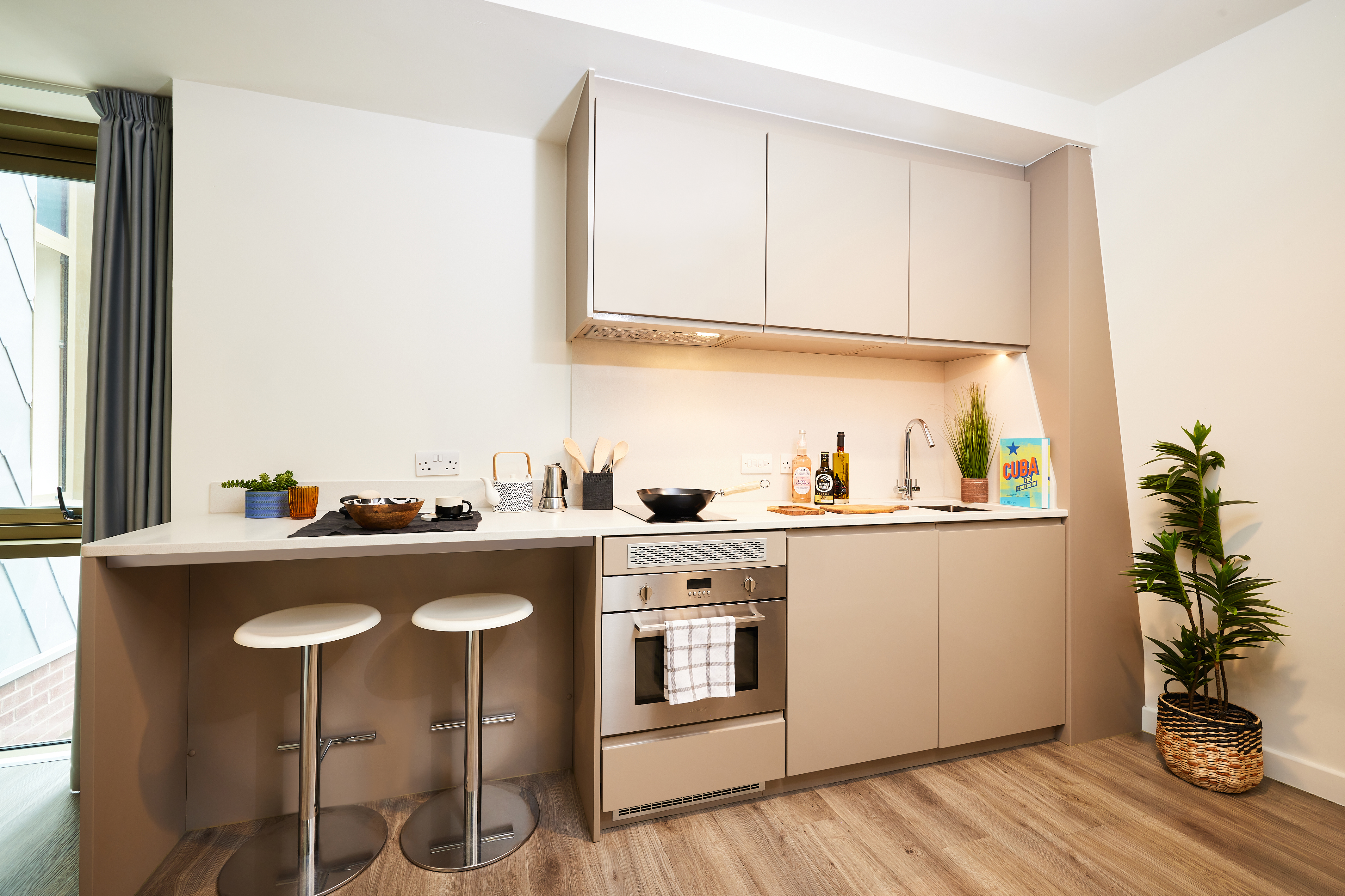 Modern kitchen with neutral cabinets, an oven, sink, and countertop. Two stools at a breakfast bar, with plants and cooking items as decor. Light wood flooring and a window with a curtain on the left.