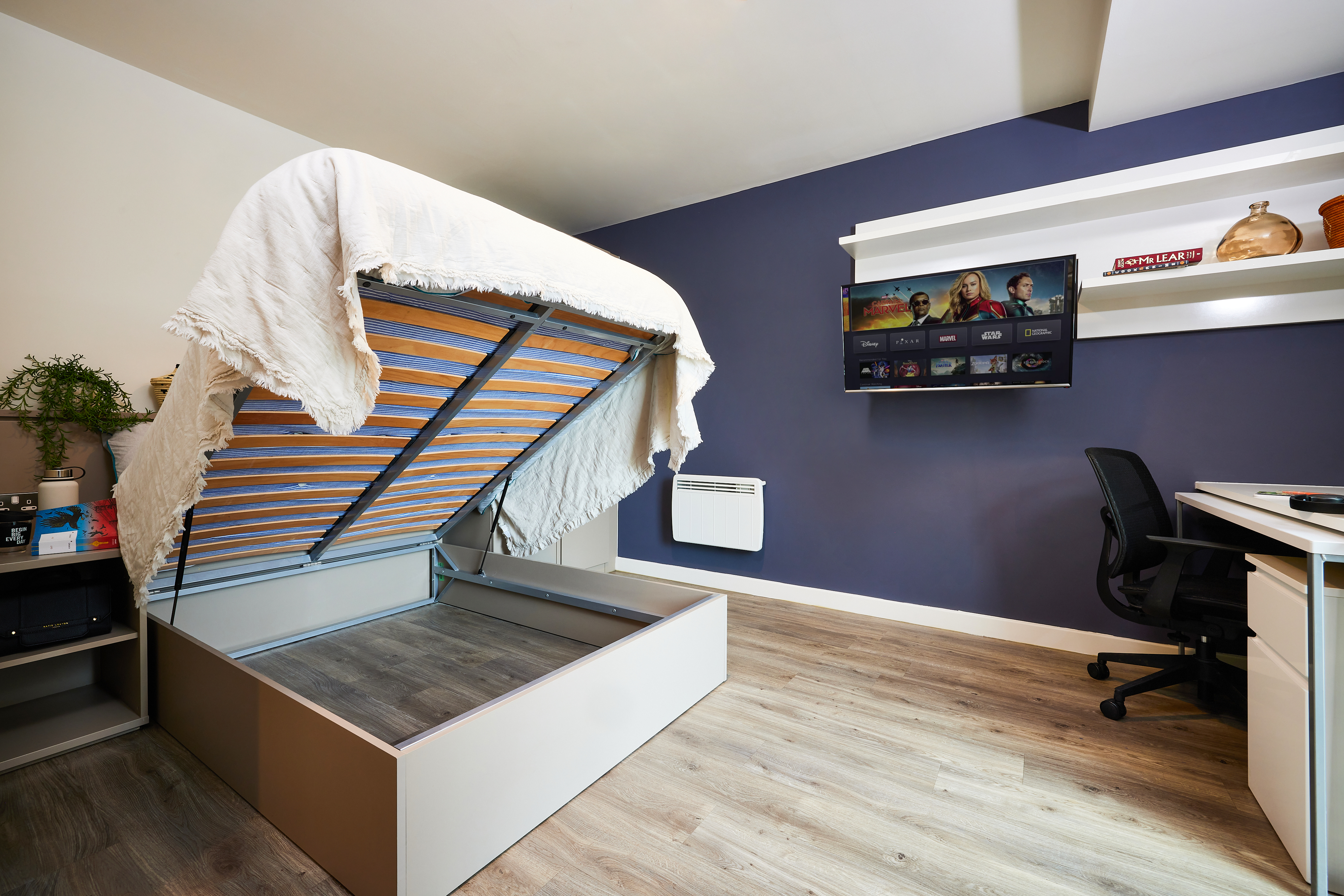 A tidy bedroom features a bed with an elevated mattress revealing storage underneath. The room has a wooden floor, a navy blue accent wall, a wall-mounted TV, a desk with a black chair, and shelves with decorative items.