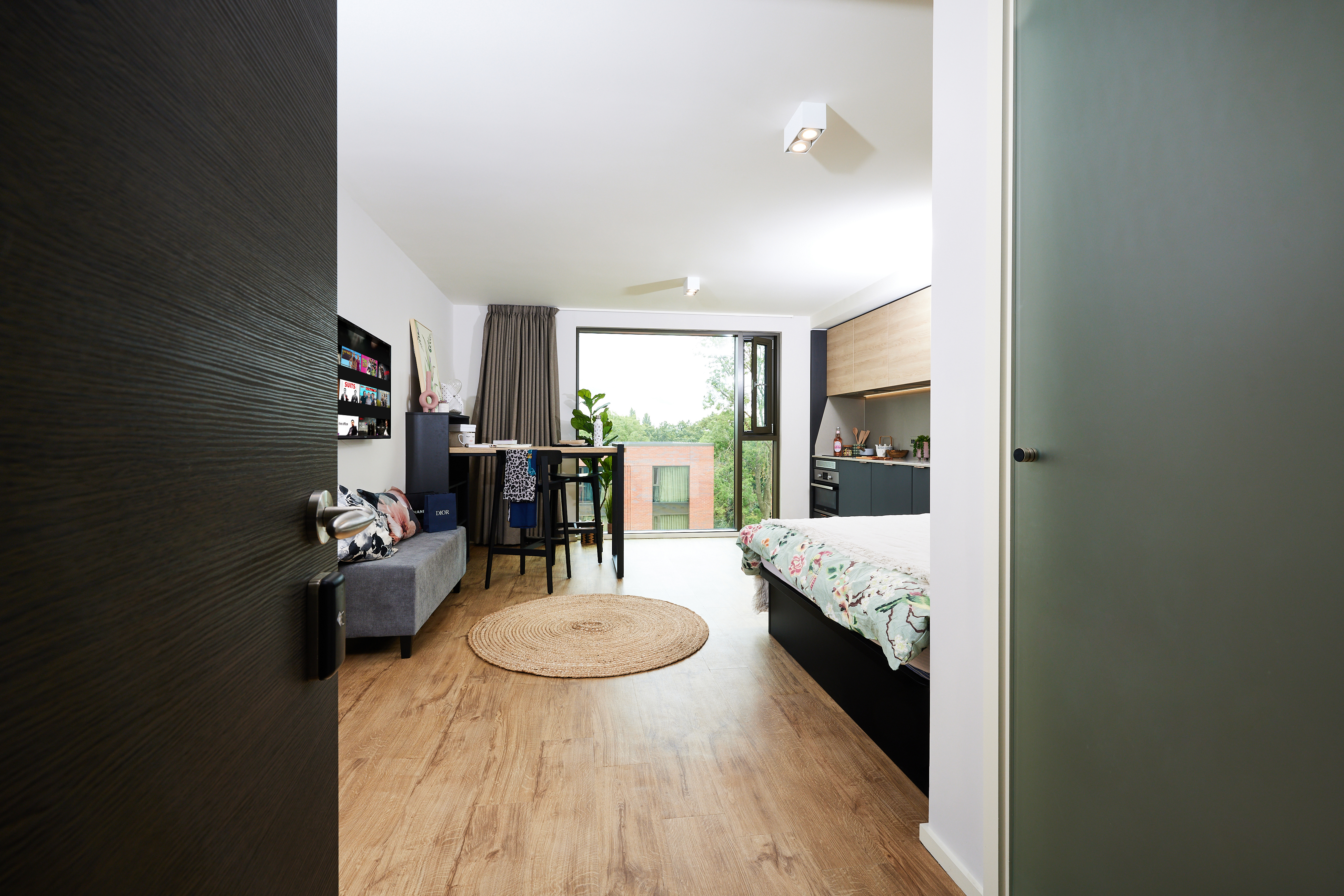 A modern studio apartment with an open door view. It features a wooden floor, a bed with floral bedding, a sofa, a desk with a chair, and a large window showing a green landscape. A round jute rug lies on the floor near the entrance.