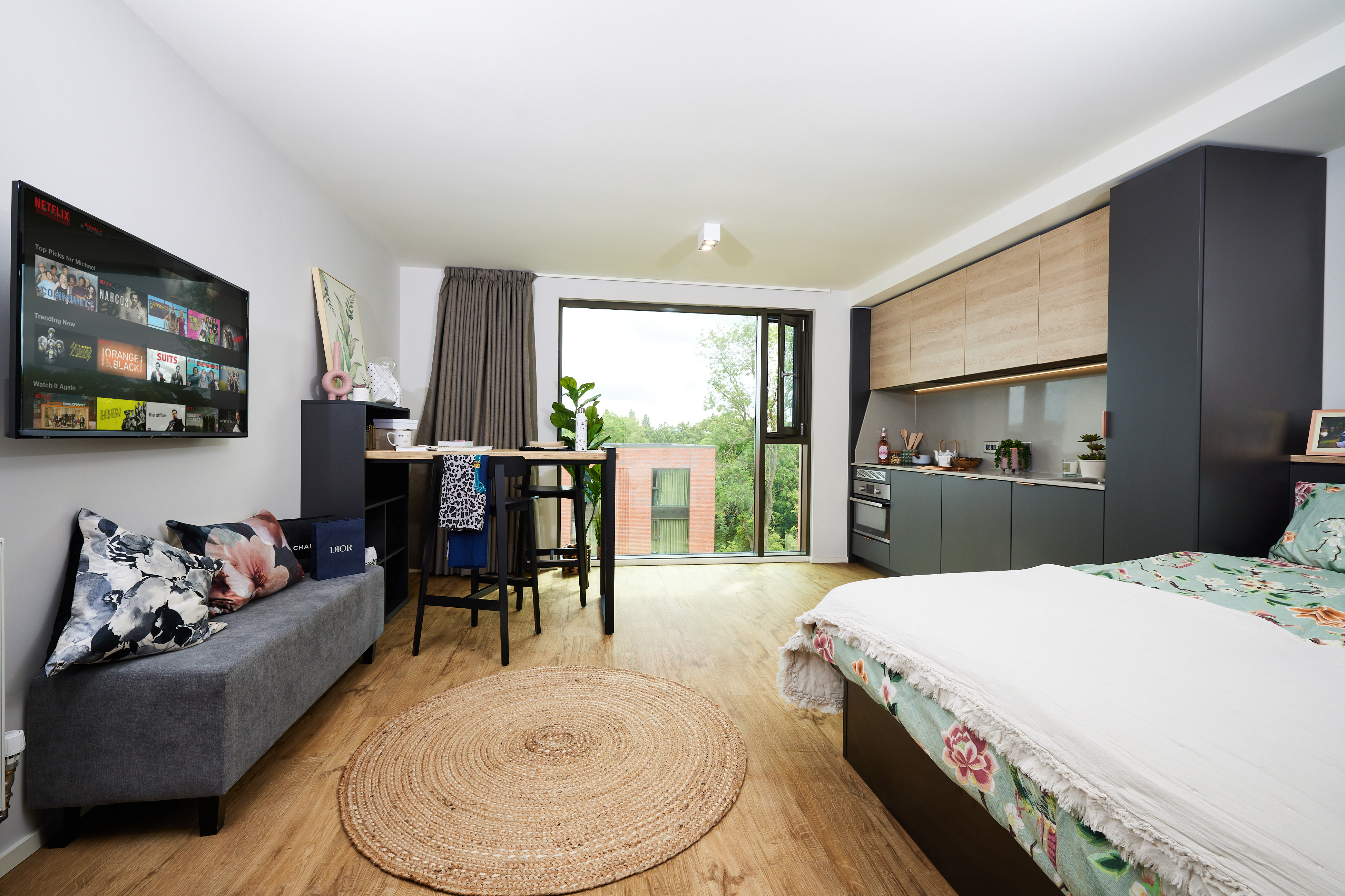 A modern studio apartment with a bed, a gray sofa, a wooden table with chairs, and a kitchen area. Large windows provide natural light, and the floor is wooden with a round woven rug. A TV is mounted on the wall.