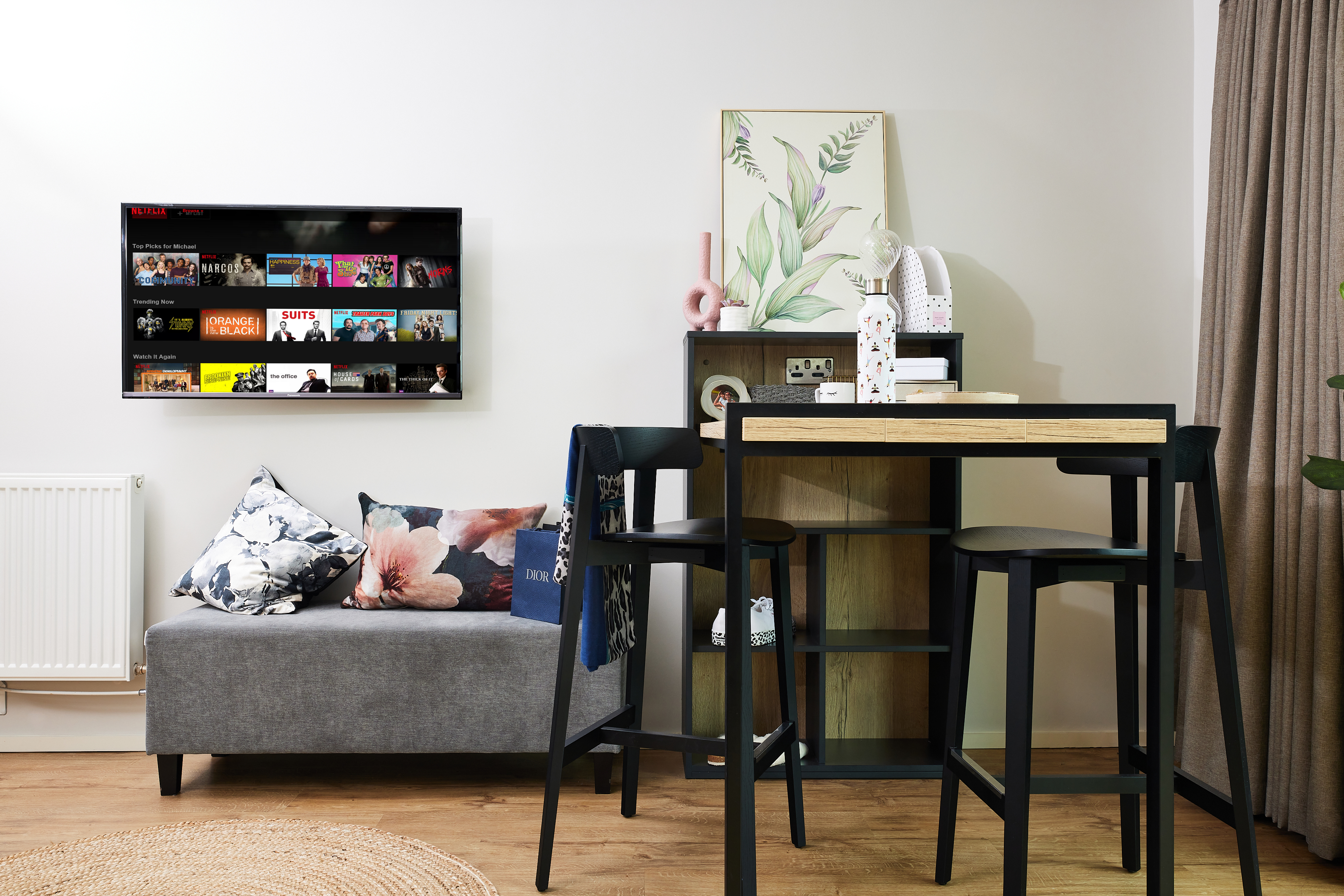 A modern living room with a small gray sofa adorned with floral cushions, a wall-mounted TV displaying streaming content, a high table with stools, decorative items, and a painting. A curtain and radiator complete the cozy setup.
