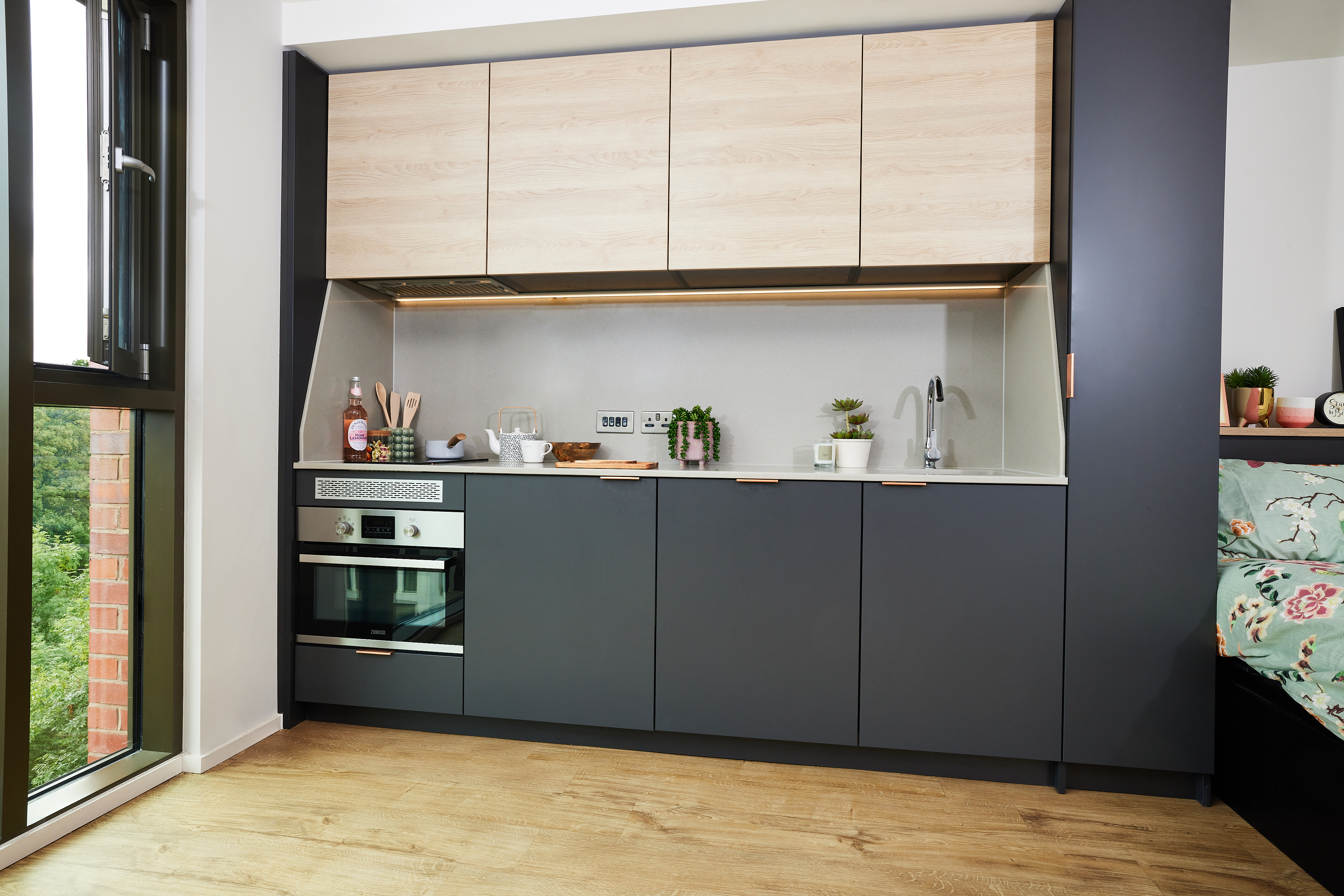 Modern kitchen with dark grey and wooden cabinets, a built-in oven, and countertop items including utensils, plants, and a kettle. A large window on the left allows natural light, illuminating the wooden floor and providing a view of greenery outside.