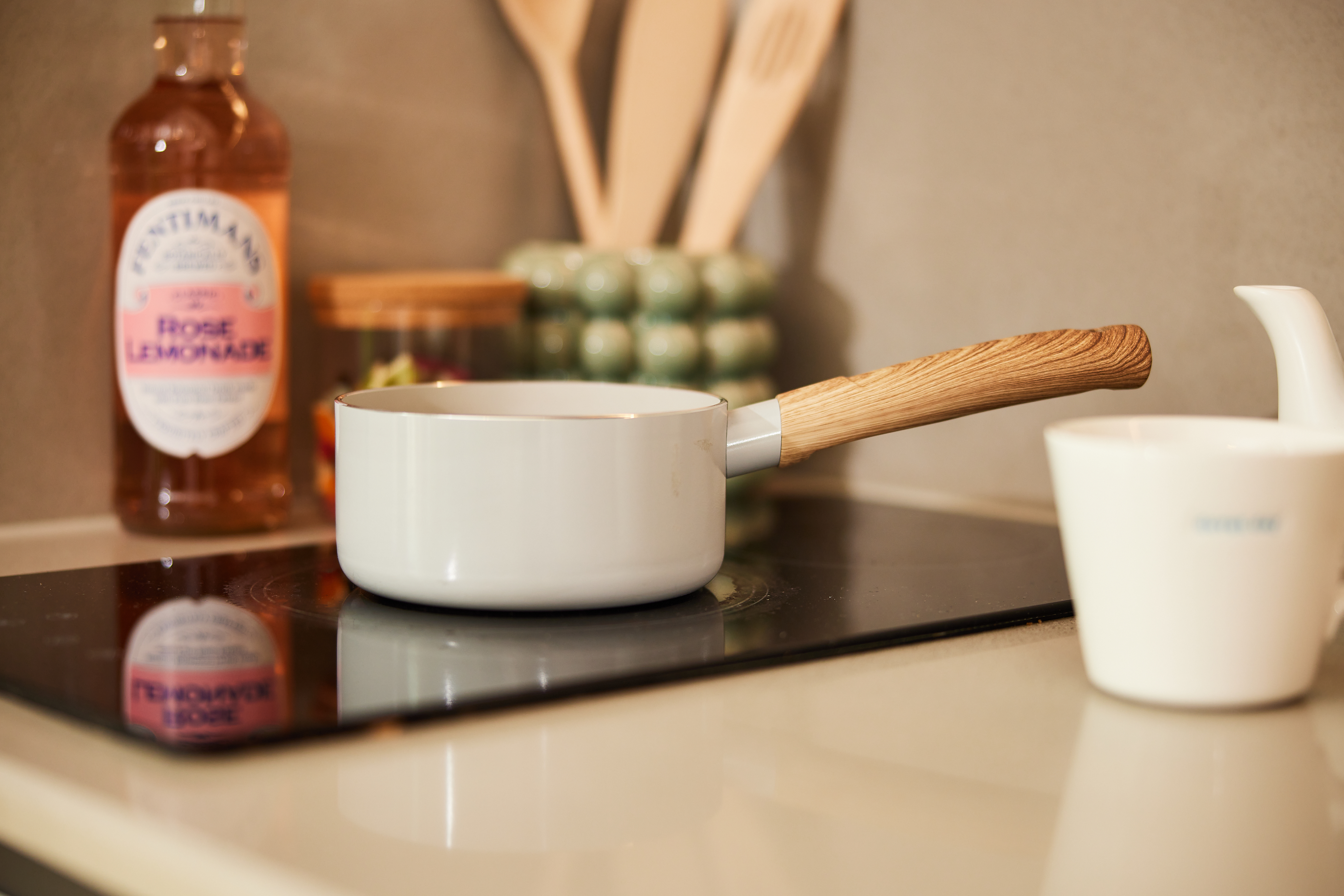 A white saucepan with a wooden handle rests on a sleek black induction stove in the heart of Portland House. Nearby, a bottle of pink lemonade, wooden utensils in a holder, and a white ceramic jug contribute to the cozy kitchen scene.