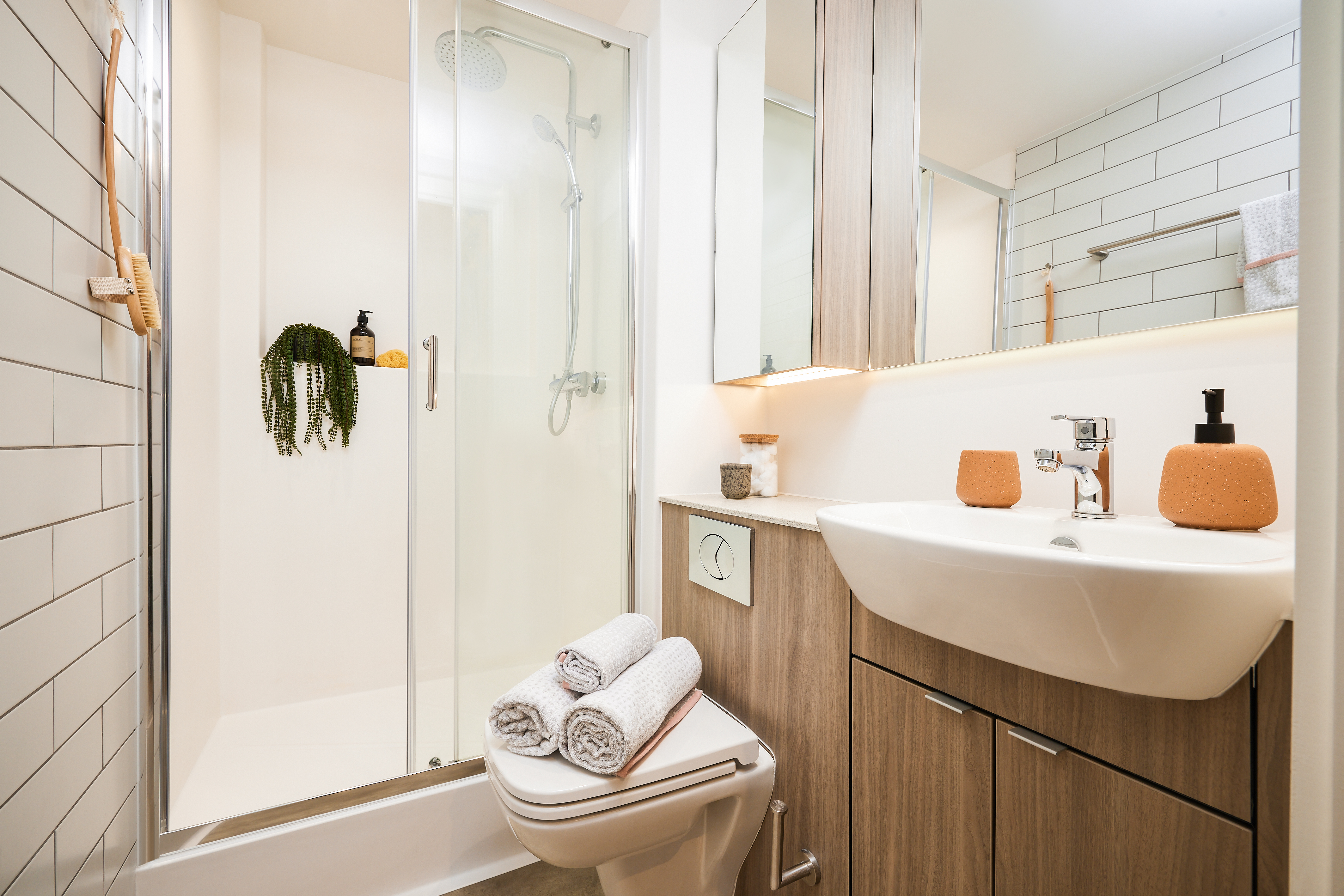 Modern bathroom with a glass-enclosed shower, white tiles, and wooden cabinets. A white sink and toilet are adorned with orange soap dispensers and rolled towels. A small plant hangs near the shower, adding a touch of greenery reminiscent of Portland's natural beauty.