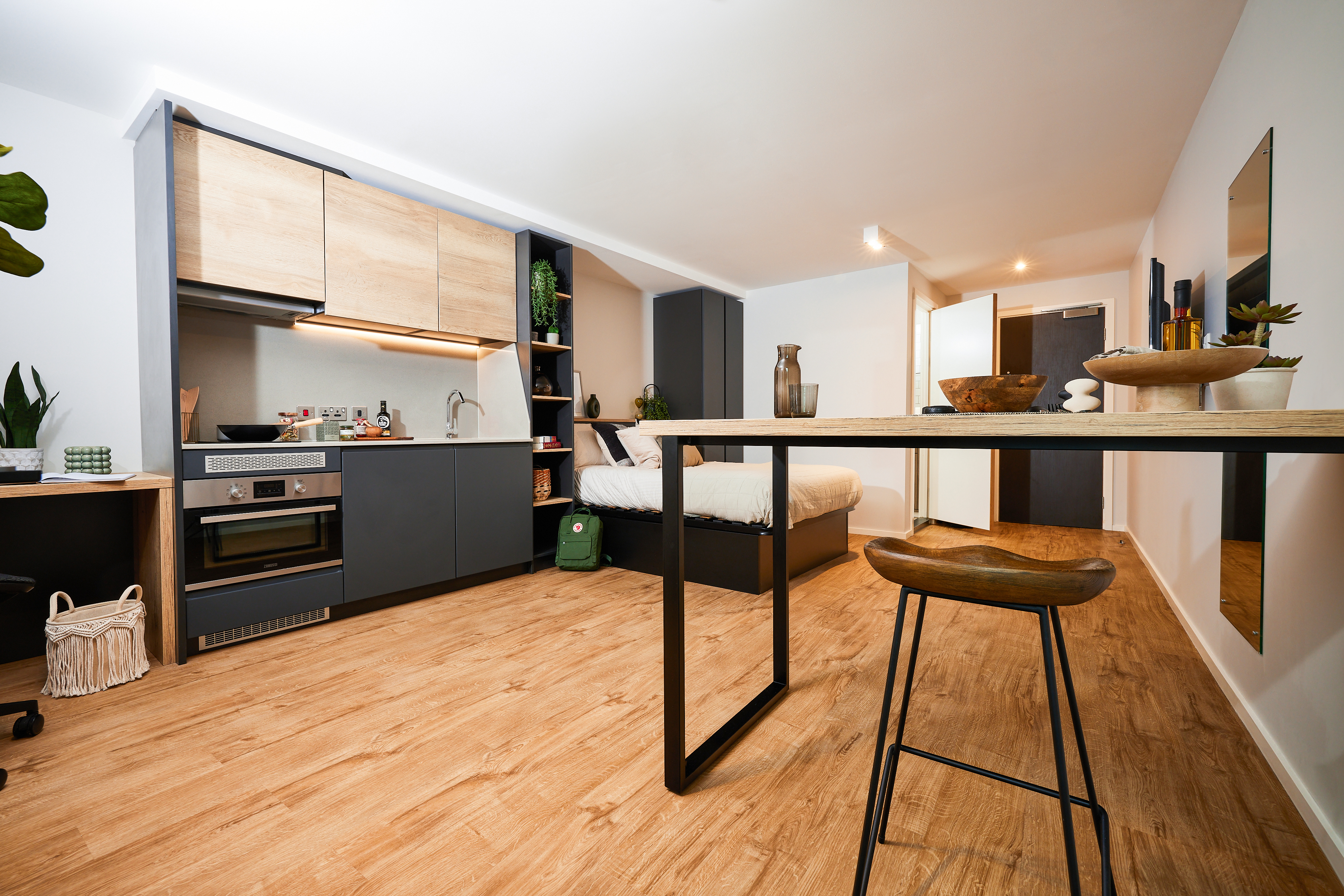 A modern studio apartment on Portland Crescent with a wooden floor, featuring a kitchenette on the left with cabinets, a microwave, and a small sink. A bed is visible in the background. A high table with a stool is in the foreground, and decor items adorn the room.