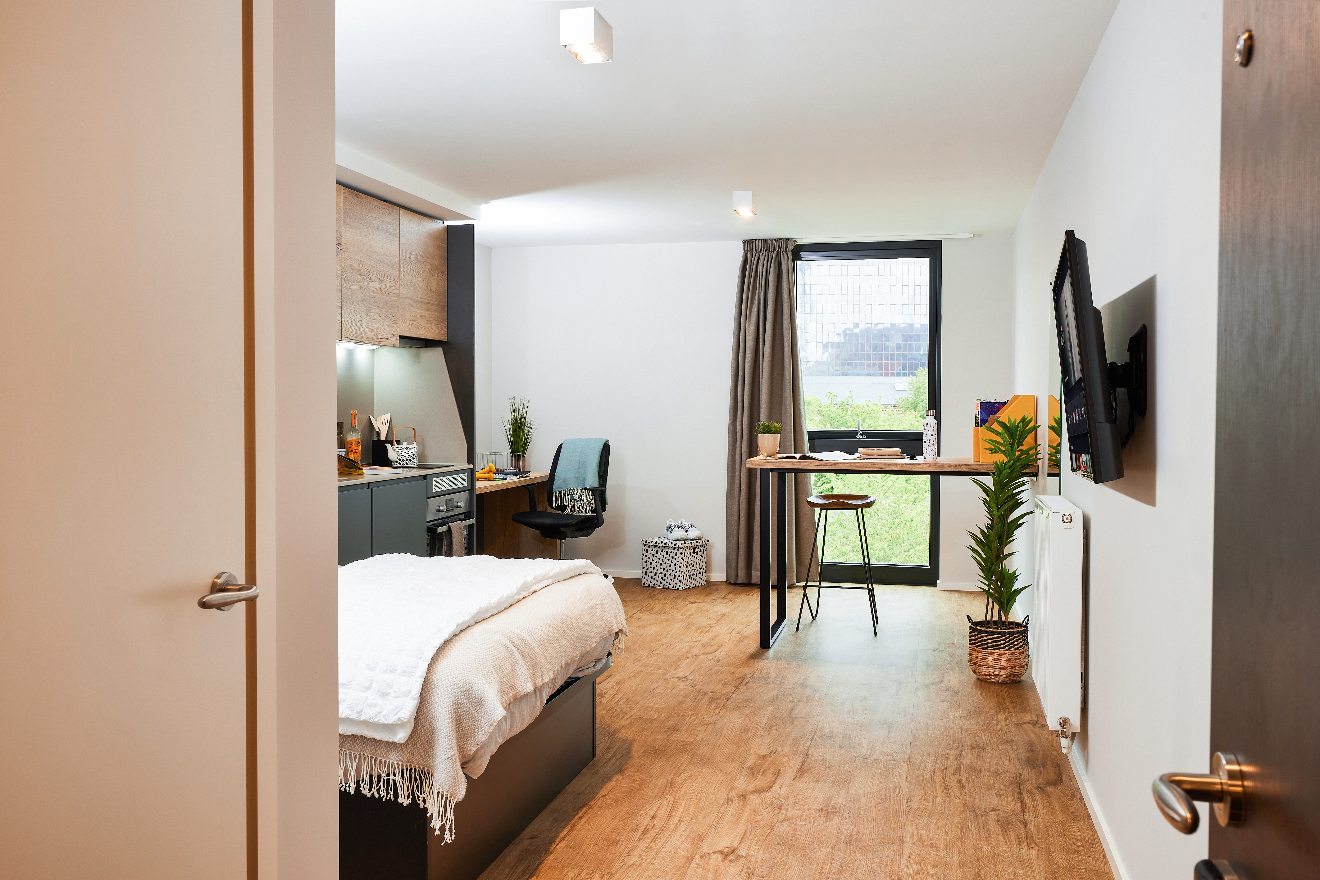 A modern studio apartment on Portland Crescent offers a bed on the left, a cozy kitchen with wooden cabinets, and a desk near a sunlit window. The space boasts wooden flooring, a wall-mounted TV, and an inspiring view of the building outside.