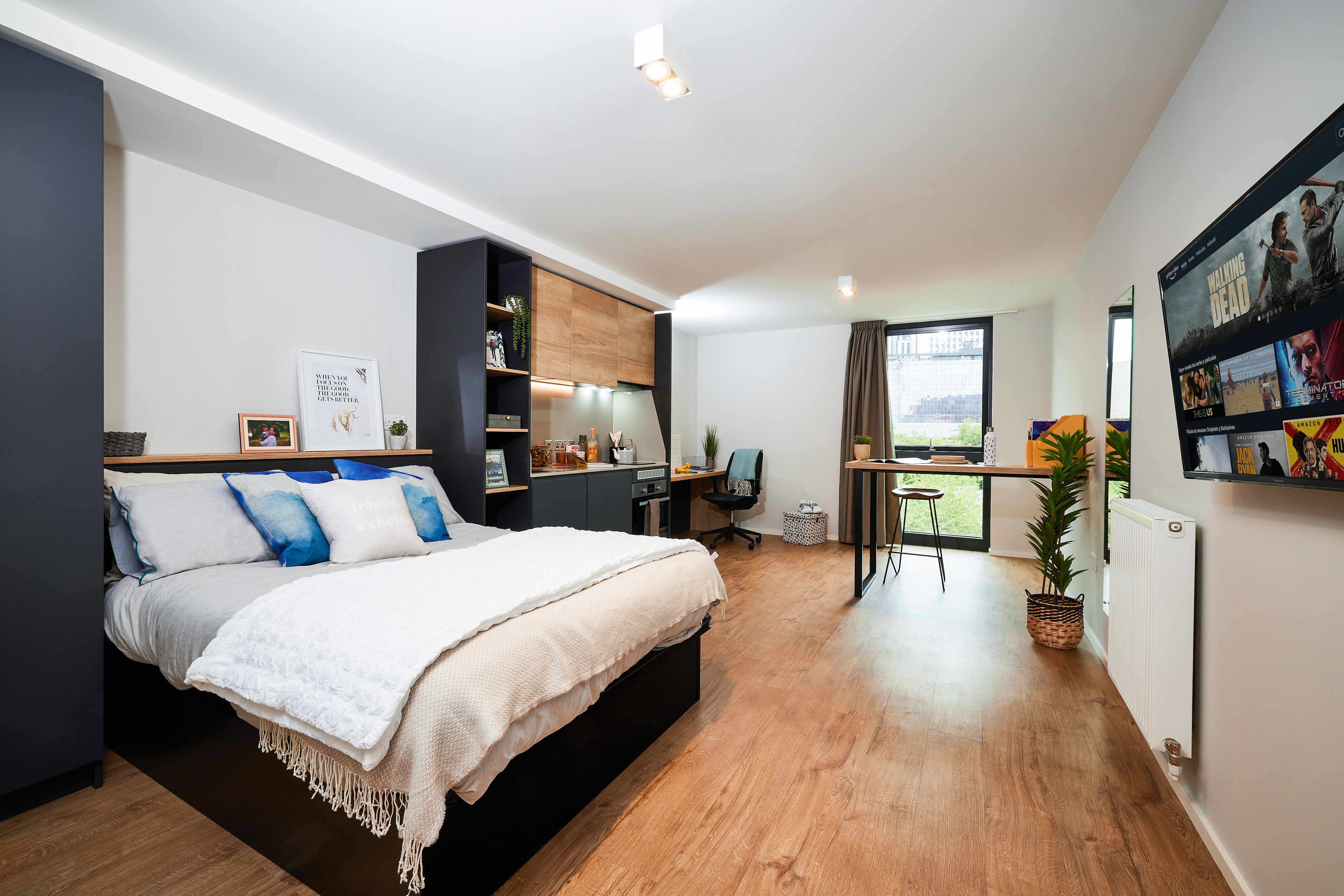A modern Portland bedroom with wood flooring features a neatly made bed with white and blue bedding. There's a desk with a chair near a window, shelves adorned with decor items, and a wall-mounted TV. The room, shaped in a gentle crescent, is brightened by abundant natural light.