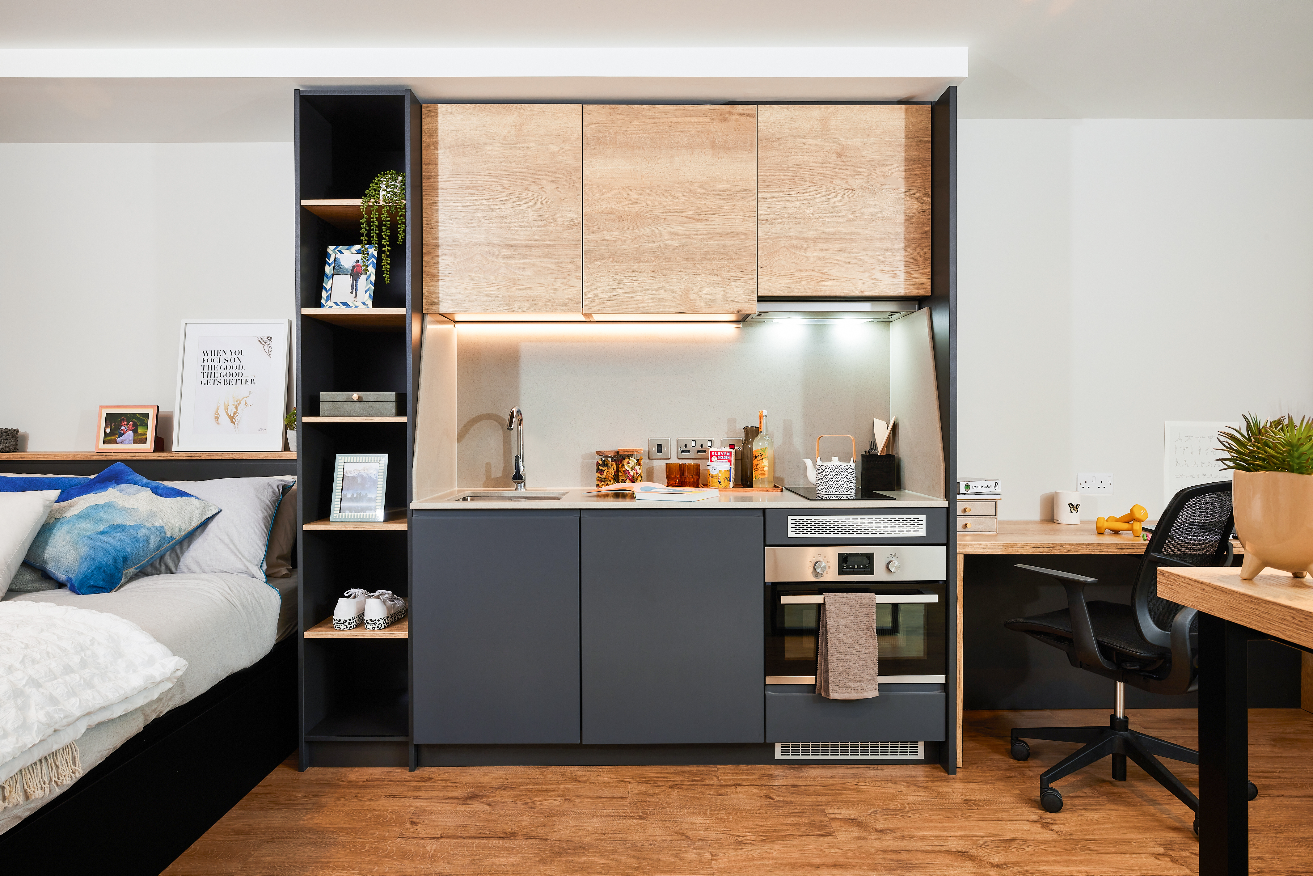 Modern studio apartment in Portland with a compact kitchen, featuring wooden cabinets, a built-in oven, and a sink. There's a cozy bed on the left and a small desk with a chair on the right. The Crescent-shaped space is bright with decorative plants and framed pictures.
