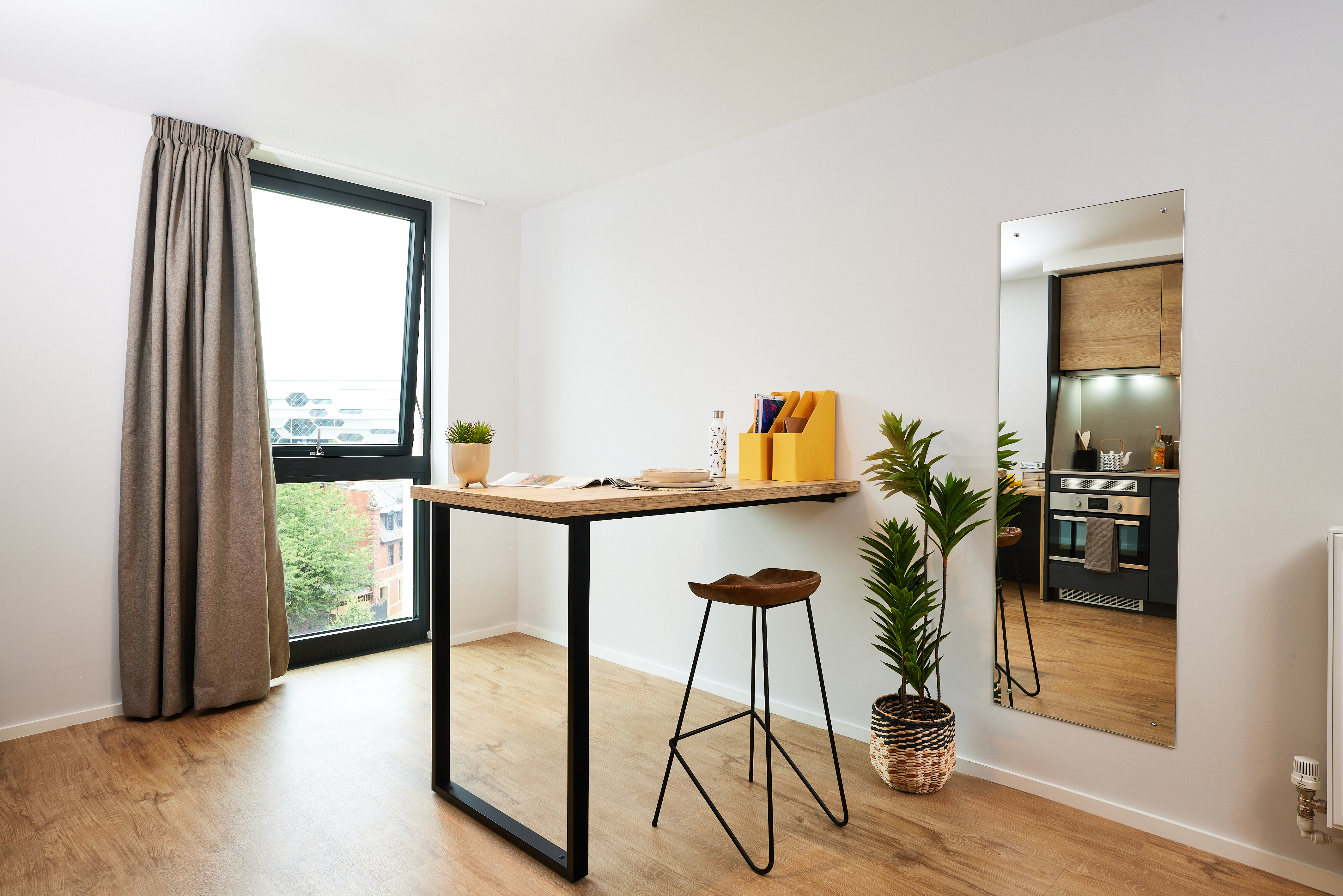 A minimalist room in Portland features a wooden desk with black metal legs and a tall mirror. A stool sits by the desk, adorned with potted plants and a yellow storage box. A large window, draped with a crescent of gray curtain, offers natural light. A kitchenette is visible in the mirror.