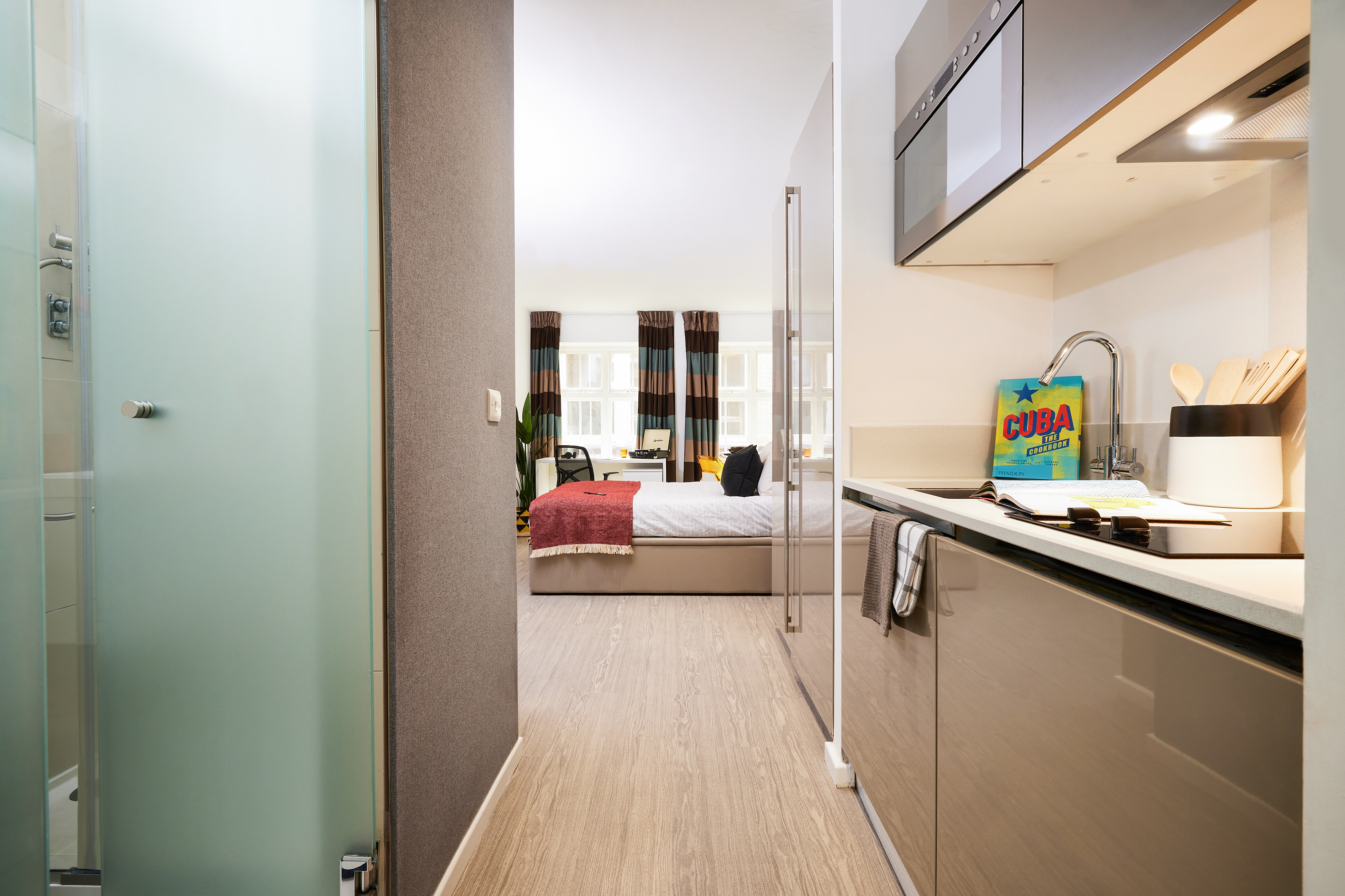 Located on Crosshall St., the modern studio apartment offers a chic kitchen with sleek cabinetry and appliances. A frosted glass door opens to reveal the bedroom area, complete with a bed draped in a red blanket and large windows offering street views.