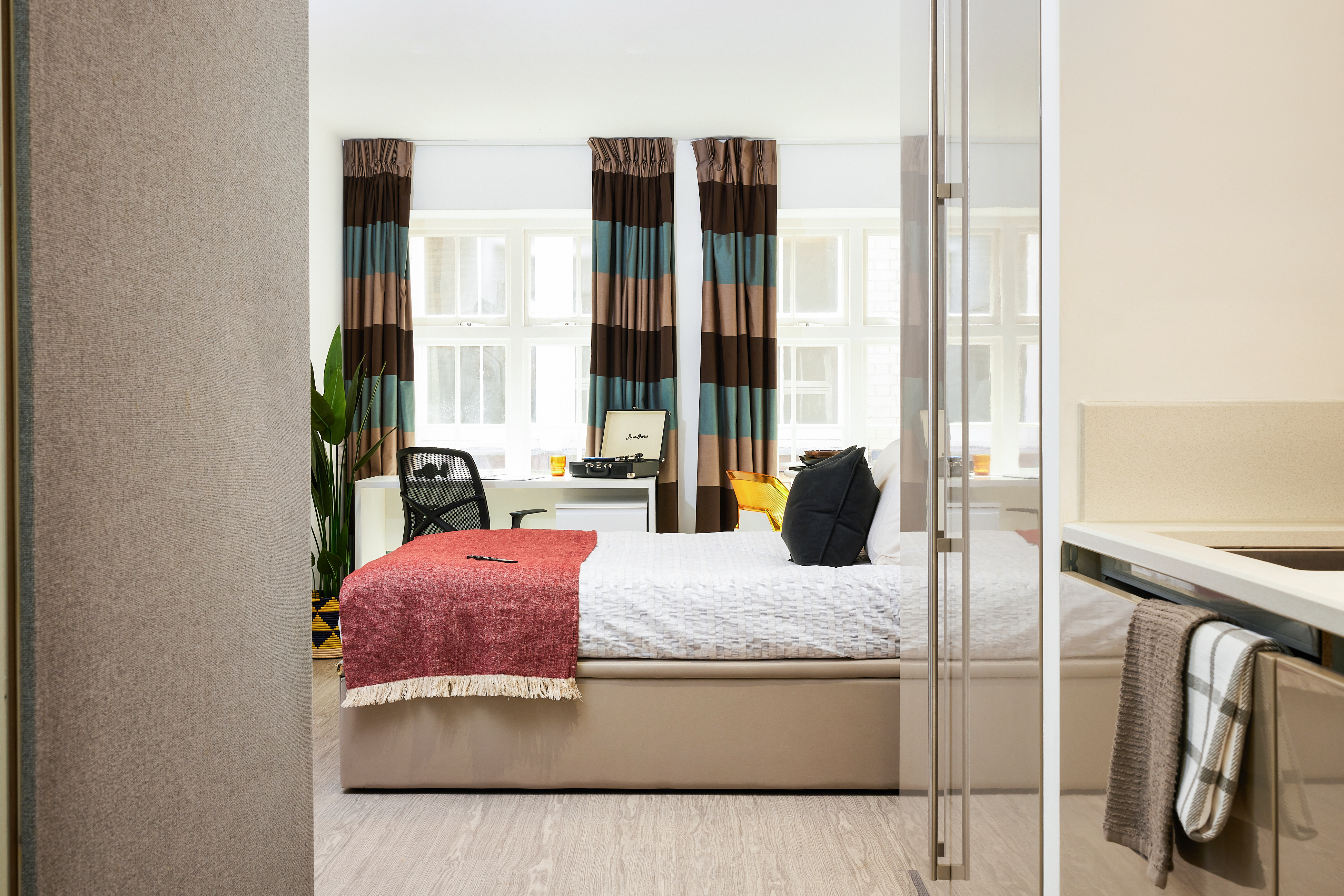 In a bright Crosshall St. bedroom, a neatly made bed with red and blue pillows invites relaxation. A red throw blanket and brown and blue curtains frame the room. By the window, a desk holds a record player beside a potted plant, offering a charming blend of style and serenity.